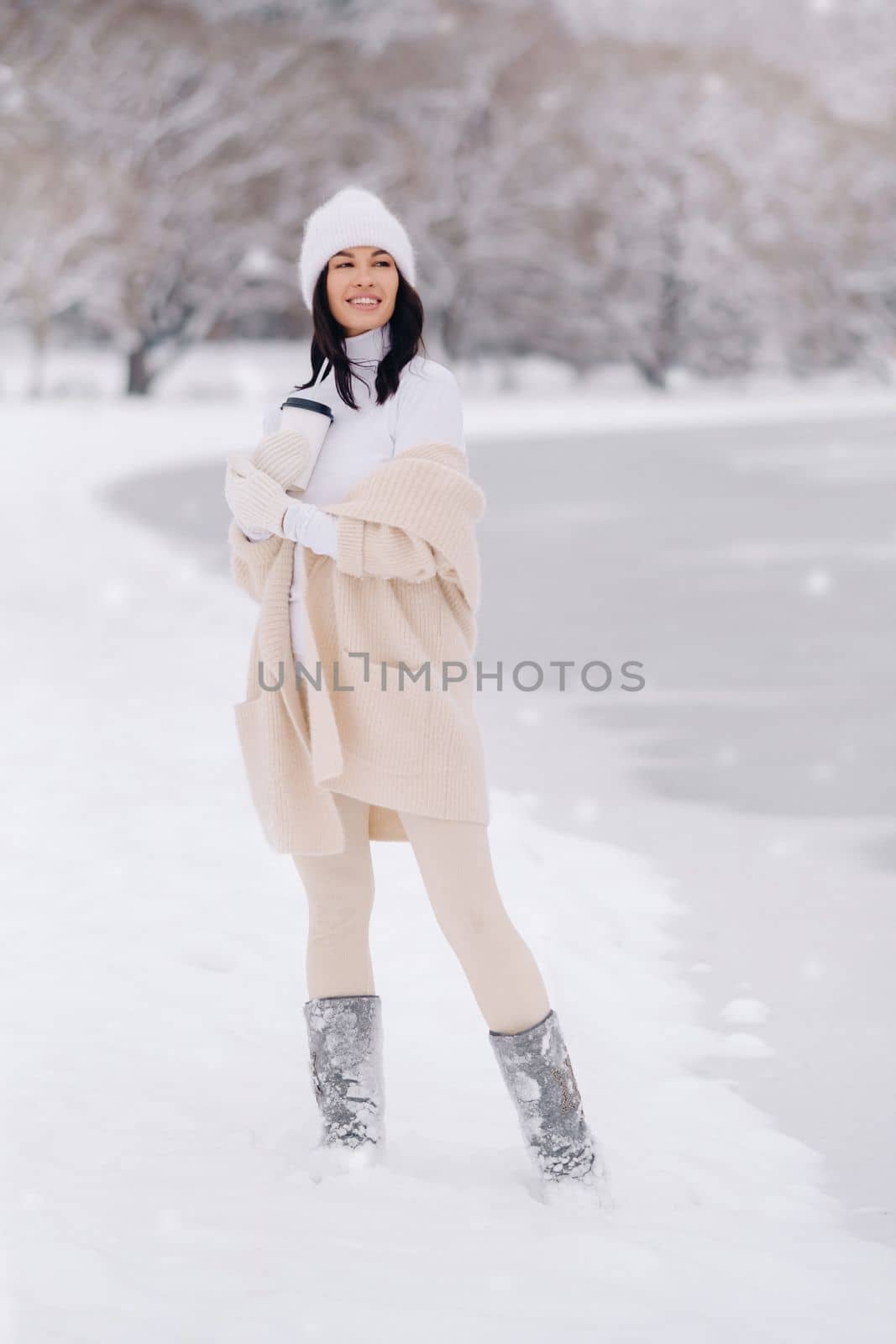 A beautiful girl in a beige cardigan and a white hat with a glass of tea enjoys a snowy embankment by the lake by Lobachad