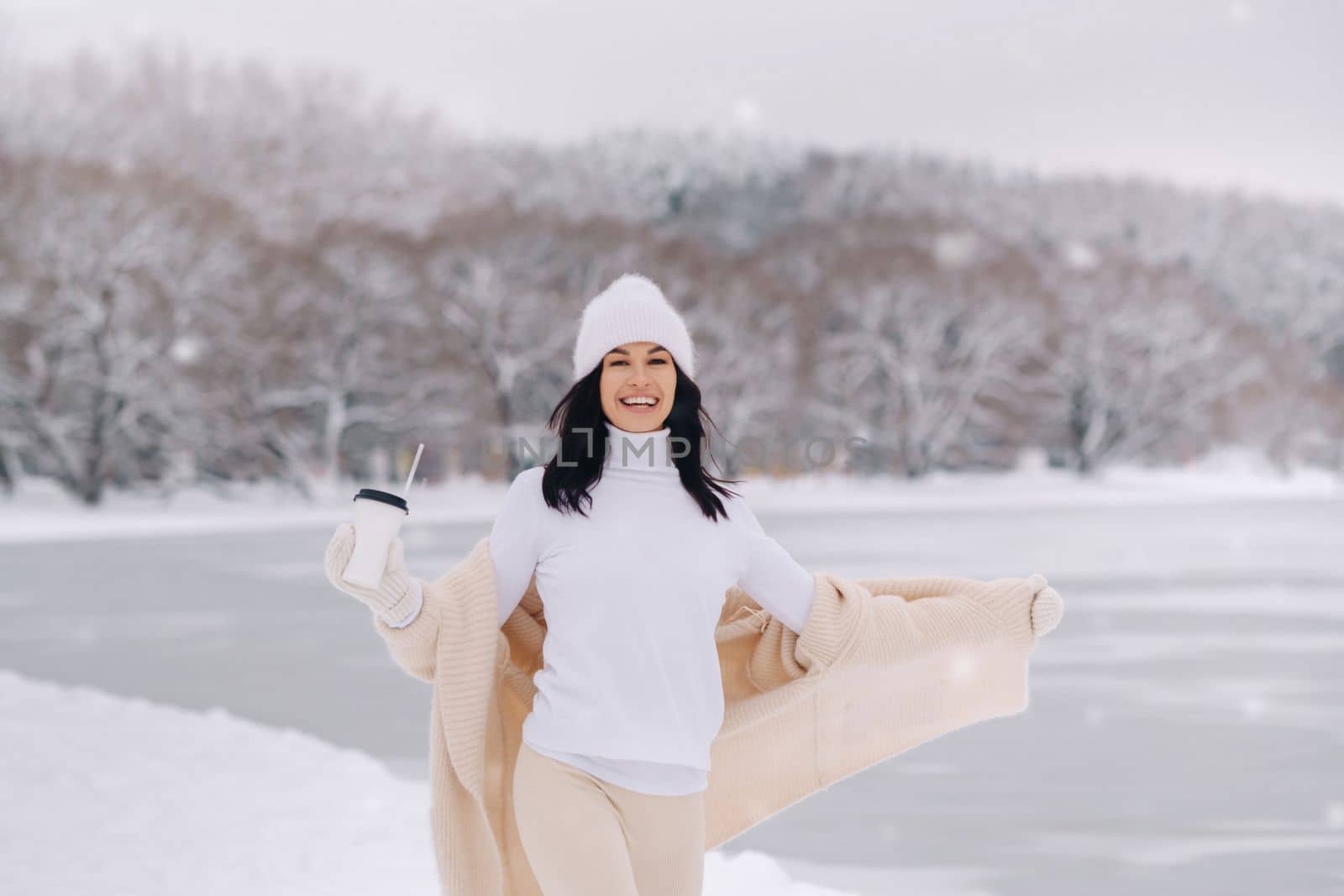 A beautiful girl in a beige cardigan and a white hat with a glass of tea enjoys a snowy embankment by the lake.