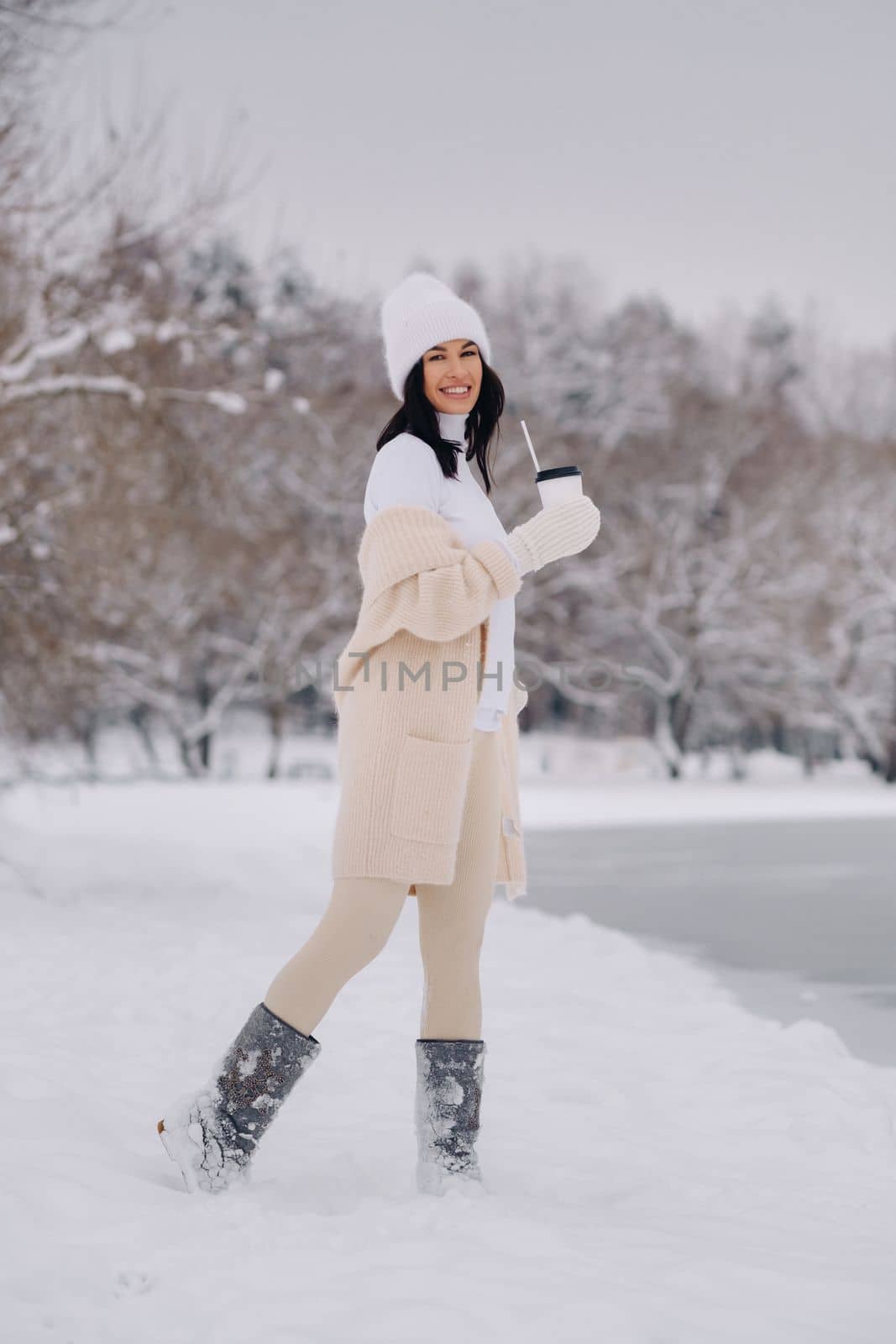 A beautiful girl in a beige cardigan and a white hat with a glass of tea enjoys a snowy embankment by the lake by Lobachad
