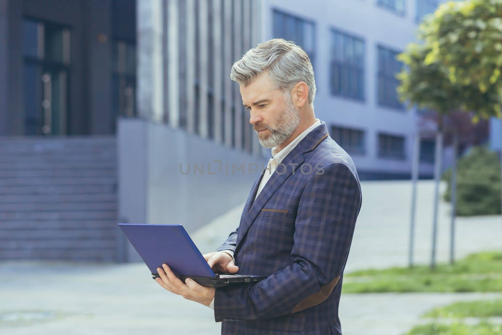 serious and concentrated gray haired boss working on laptop while standing outside modern office building, mature boss in business suit typing on netbook keyboard, senior successful businessman.