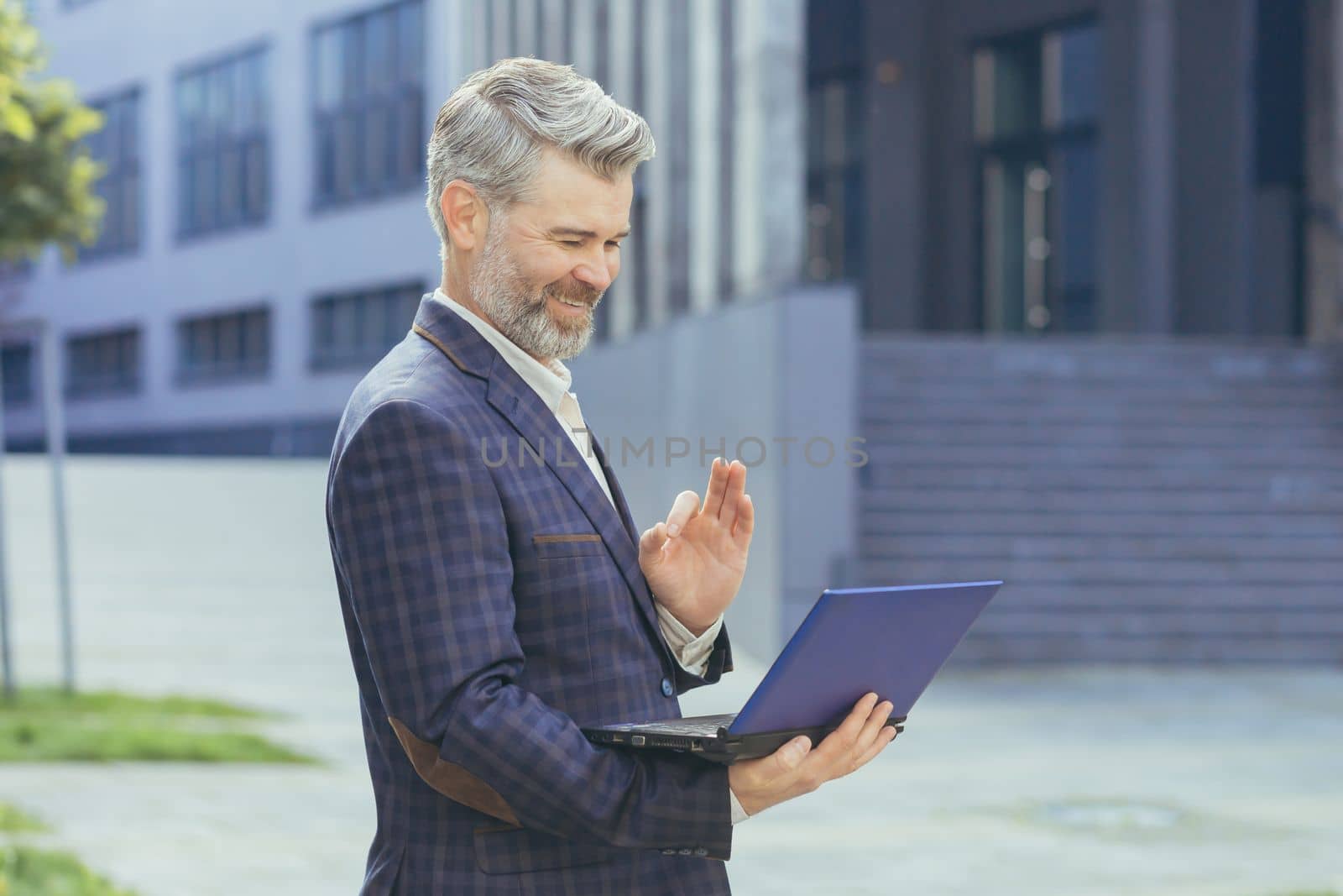 Mature boss talking standing on video call, senior gray-haired businessman holding netbook looking at web camera and explaining discussing, man in business suit outside office building with laptop.