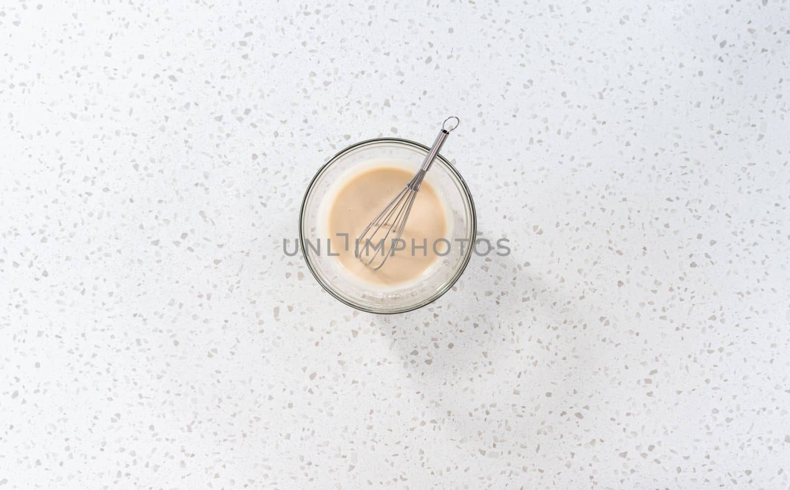 Flat lay. Mixing ingredients with a hand whisk in a glass mixing bowl to make the vanilla glaze.