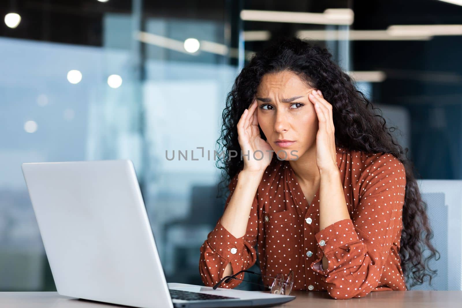 Overtired hispanic business woman working inside modern office, office worker with laptop having severe headache.