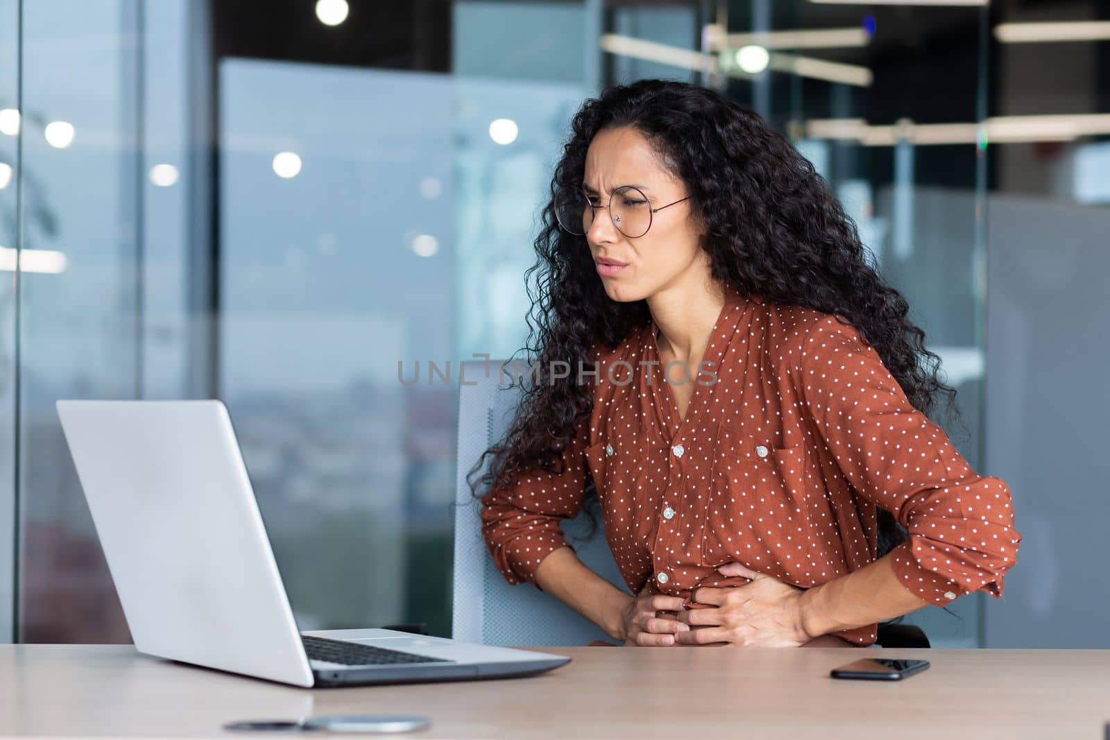 Sick business woman working inside office using laptop while sitting at table, Hispanic woman has severe stomach pain.