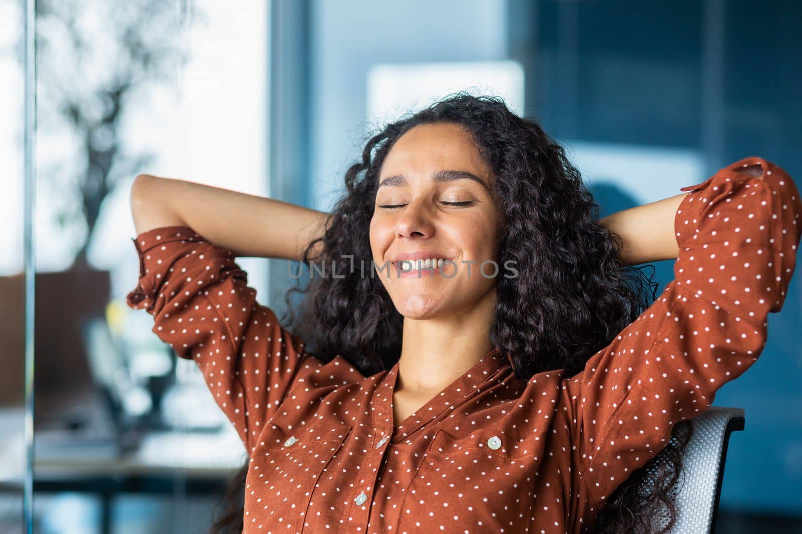 Happy and smiling successful hispanic business woman finished work satisfied with work result and achievement, hands behind head and smiling closeup.