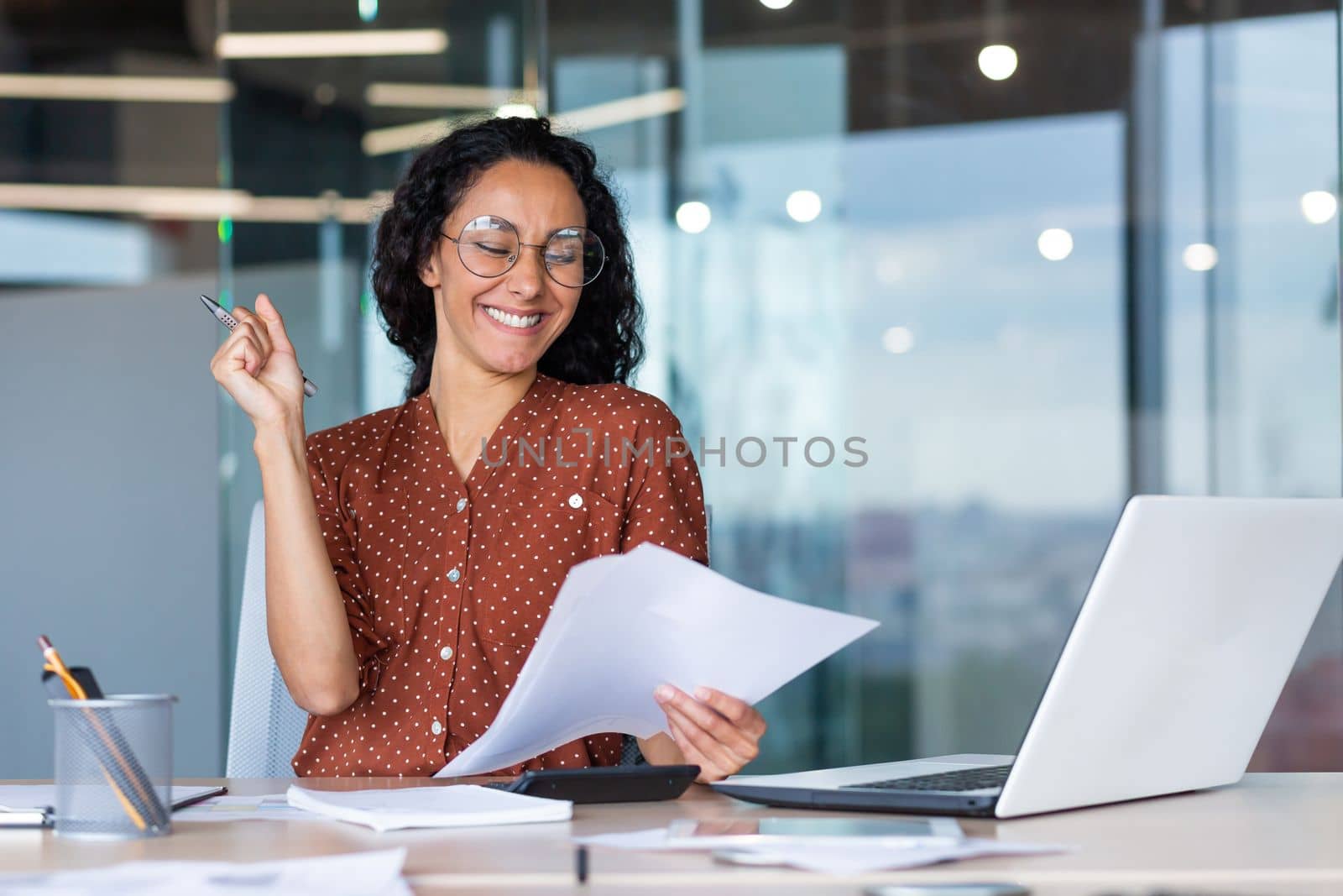 Businesswoman celebrating victory and successful achievement, Hispanic woman behind paper work reading documents with positive results reports, woman working on laptop inside office at table sitting.