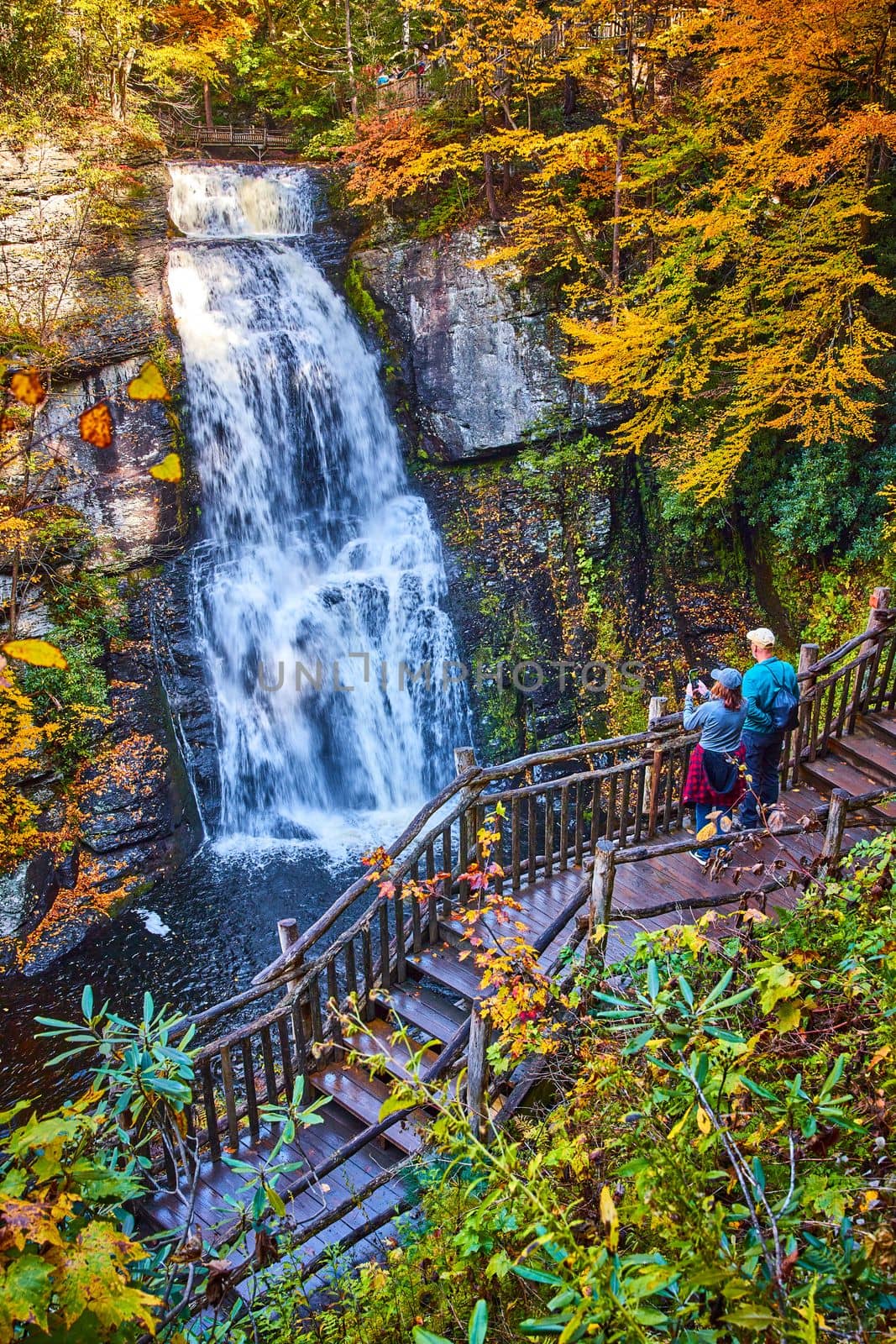 Image of Couple enjoying view from boardwalk of huge stunning waterfall over cliffs surrounded by fall foliage