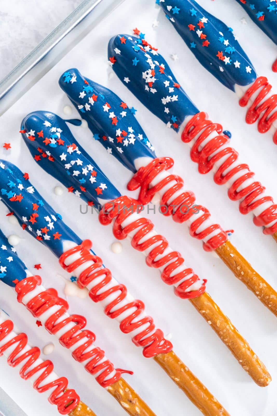 Homemade chocolate-covered pretzel rods decorated like the American flag drying on a baking sheet lined with parchment paper.