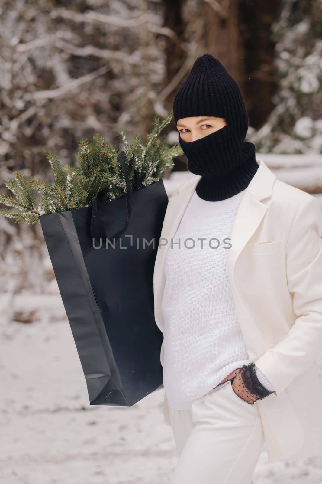 A girl in a white suit and balaclava with a package of Christmas trees in the winter forest on New Year's Eve.New Year's concept by Lobachad