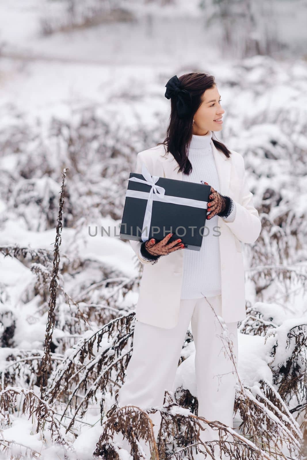 A stylish woman with a white suit with a New Year's gift in her hands in a winter forest. A girl in nature in a snowy forest with a gift box by Lobachad