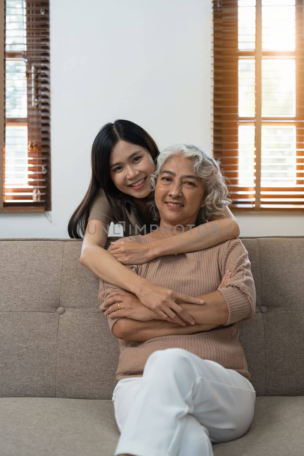Loving adult daughter hugging older mother on couch at home, family enjoying tender moment together, young woman and mature mum or grandmother looking at each other, two generations.
