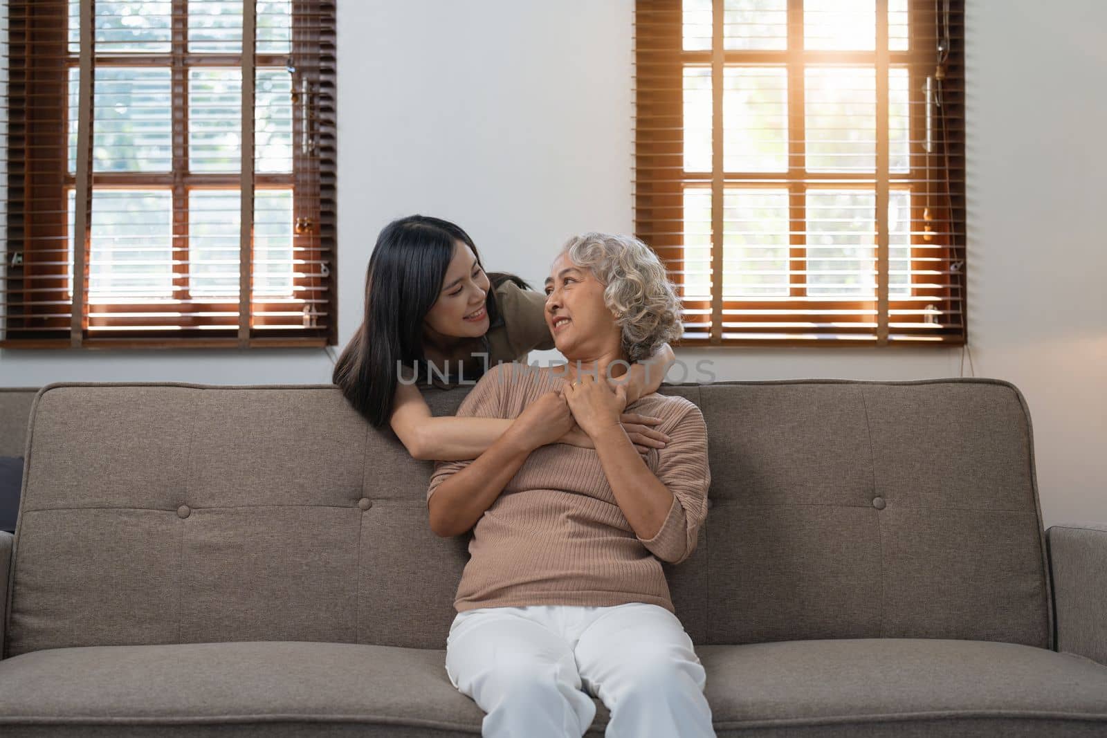 Loving adult daughter hugging older mother on couch at home, family enjoying tender moment together, young woman and mature mum or grandmother looking at each other, two generations by itchaznong