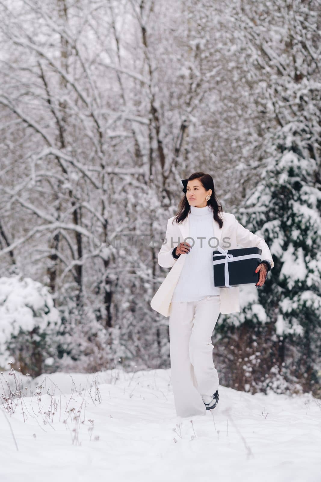 A stylish woman with a white suit with a New Year's gift in her hands in a winter forest. A girl in nature in a snowy forest with a gift box by Lobachad