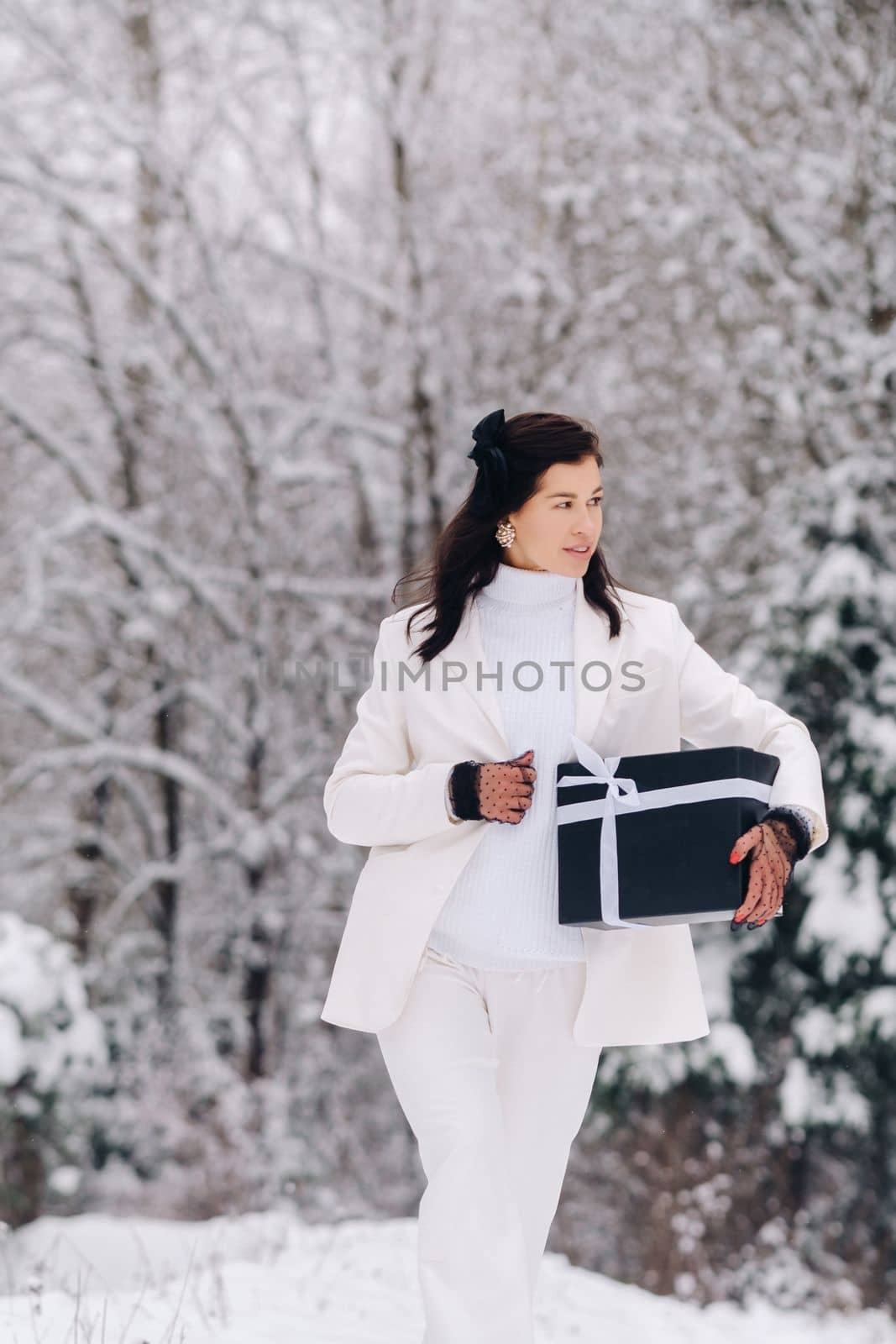 A stylish woman with a white suit with a New Year's gift in her hands in a winter forest. A girl in nature in a snowy forest with a gift box.