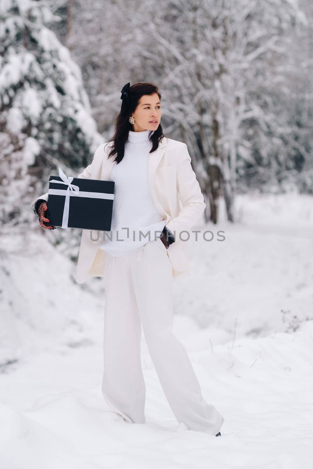 A stylish woman with a white suit with a New Year's gift in her hands in a winter forest. A girl in nature in a snowy forest with a gift box.