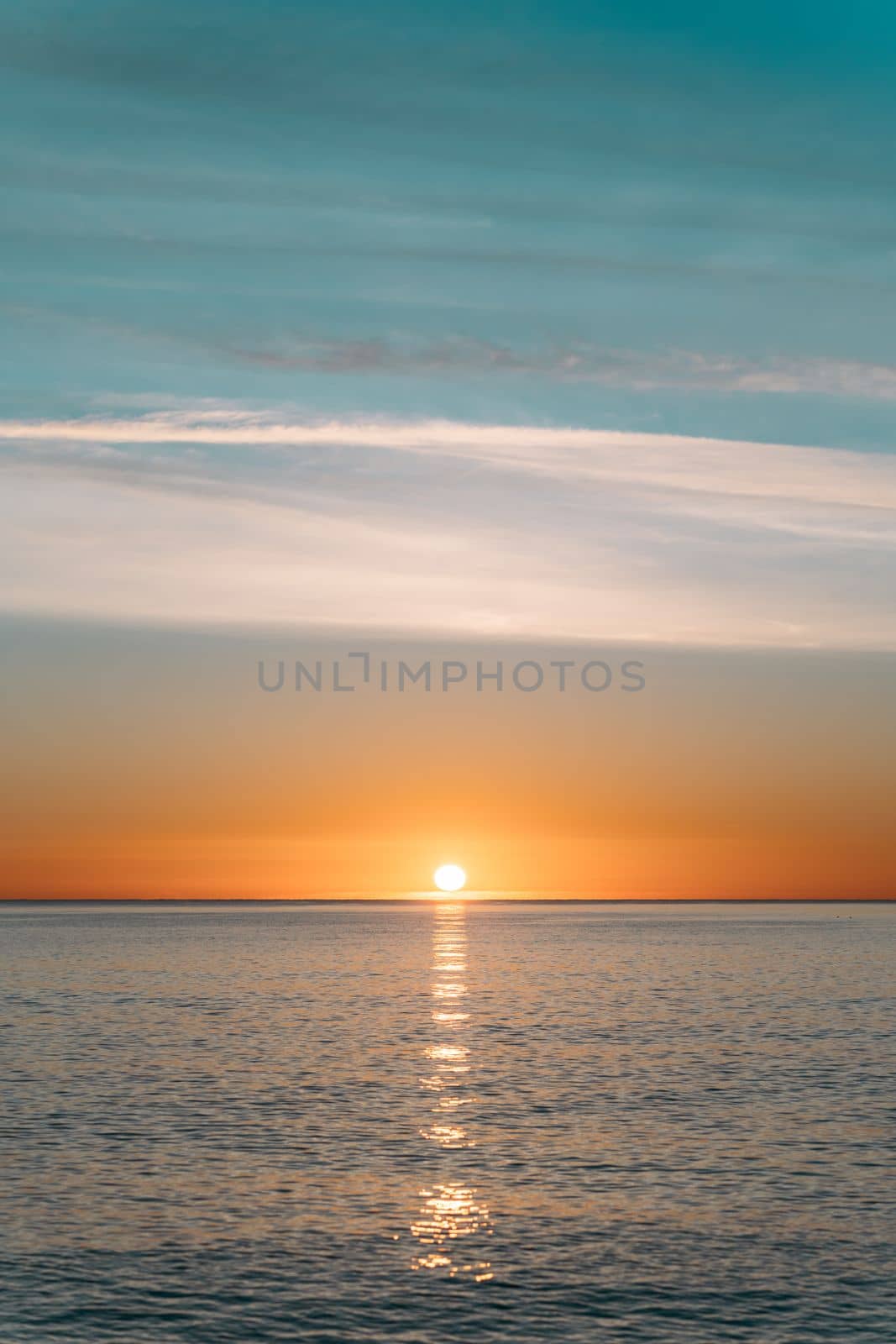An amazing sunset with bright orange hues against a blue cloudless sky. A ray of sun casts a bright glare on the calm water surface of the ocean. The perfect evening in nature. by apavlin
