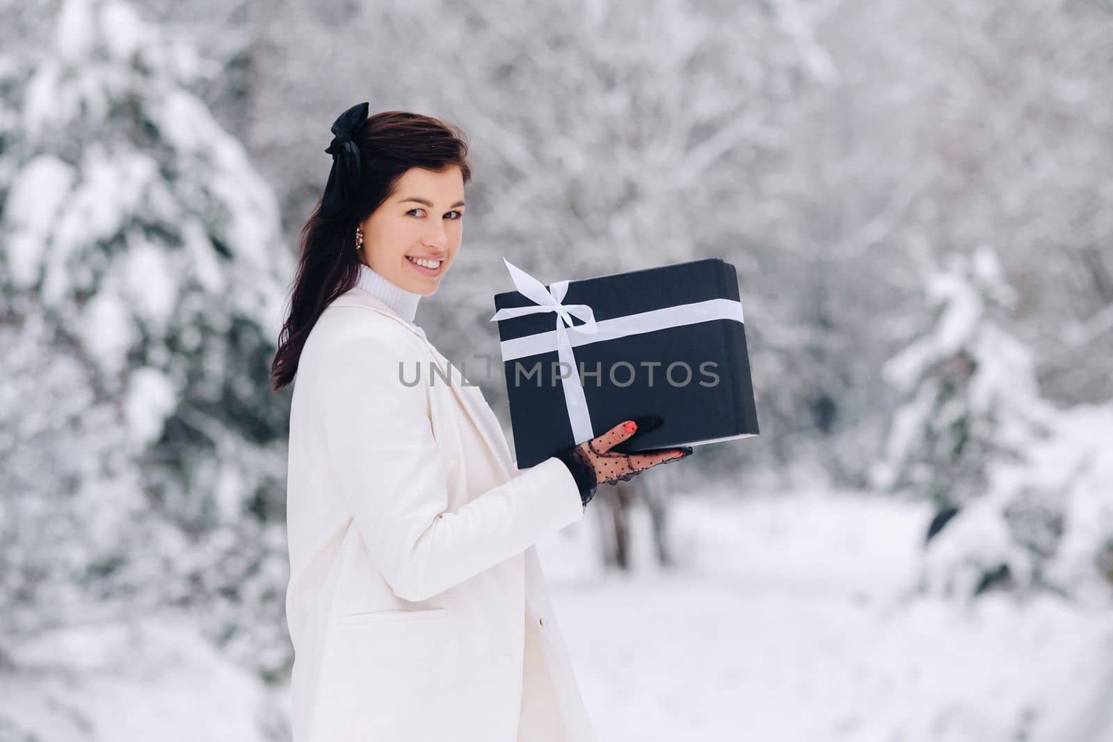 A stylish woman with a white suit with a New Year's gift in her hands in a winter forest. A girl in nature in a snowy forest with a gift box.