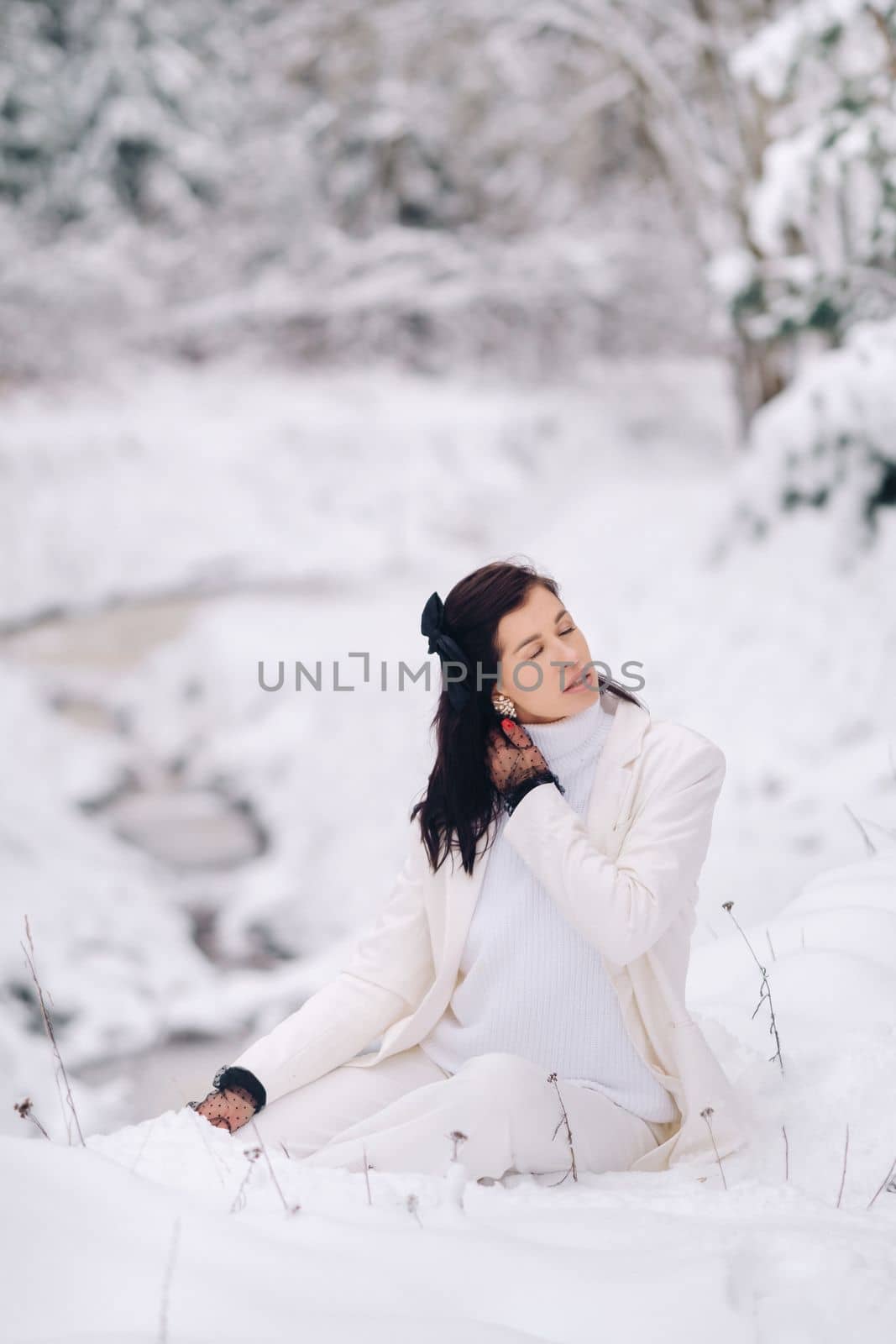 Portrait of a stylish woman in a white suit with elegant gloves sitting in nature in winter by Lobachad