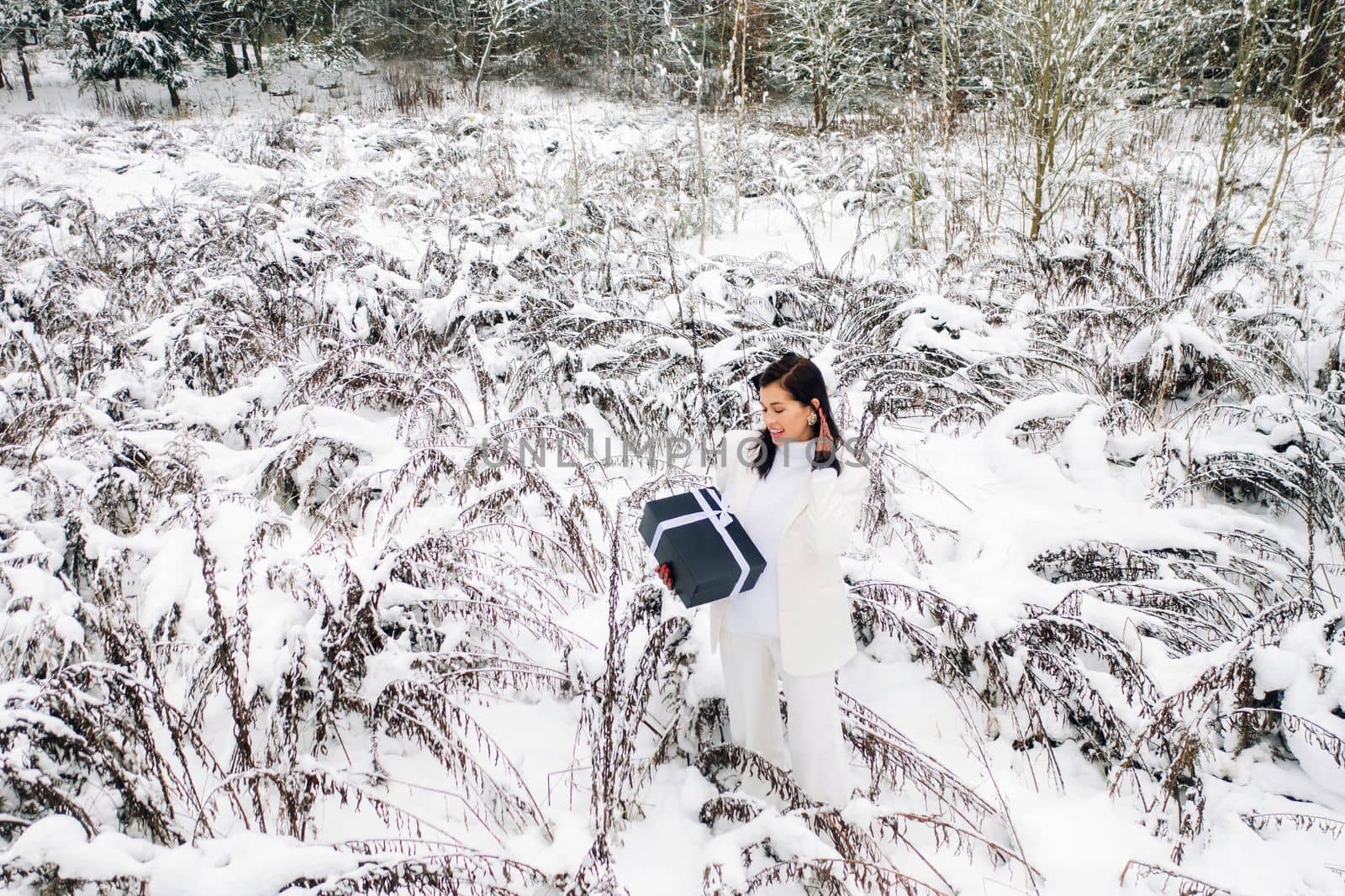 A stylish woman with a white suit with a New Year's gift in her hands in a winter forest. A girl in nature in a snowy forest with a gift box by Lobachad