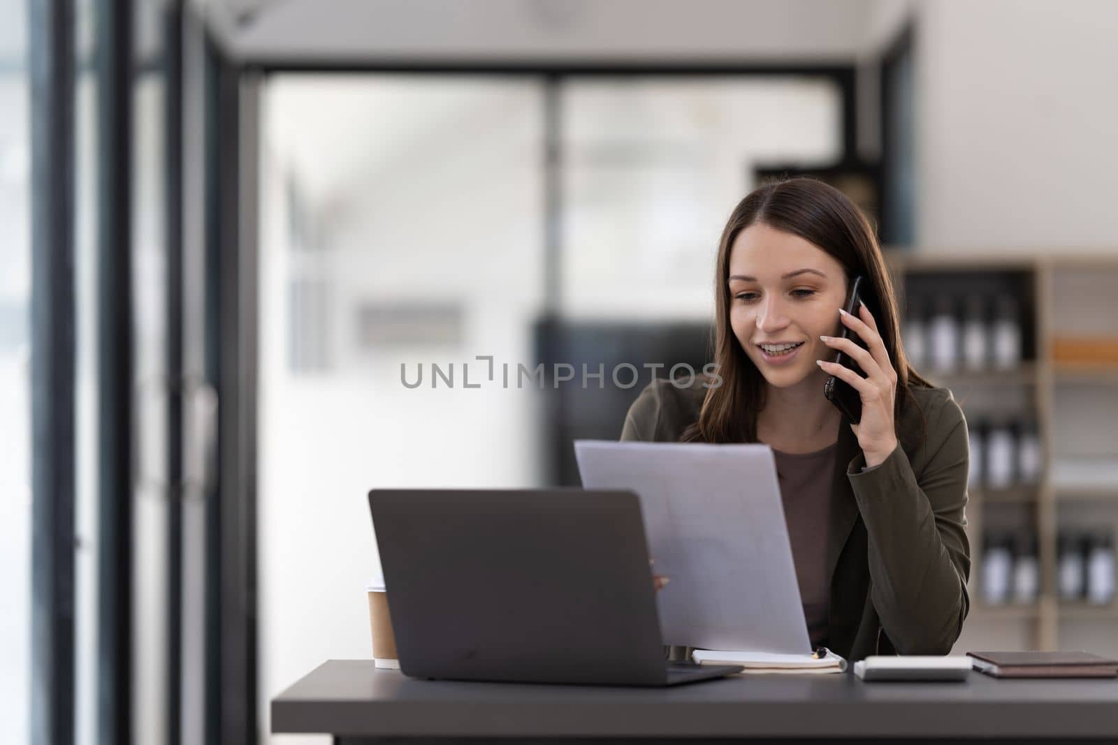 Happy smiling business woman working on laptop at office, using smart phone. Businesswoman sitting at her working place.