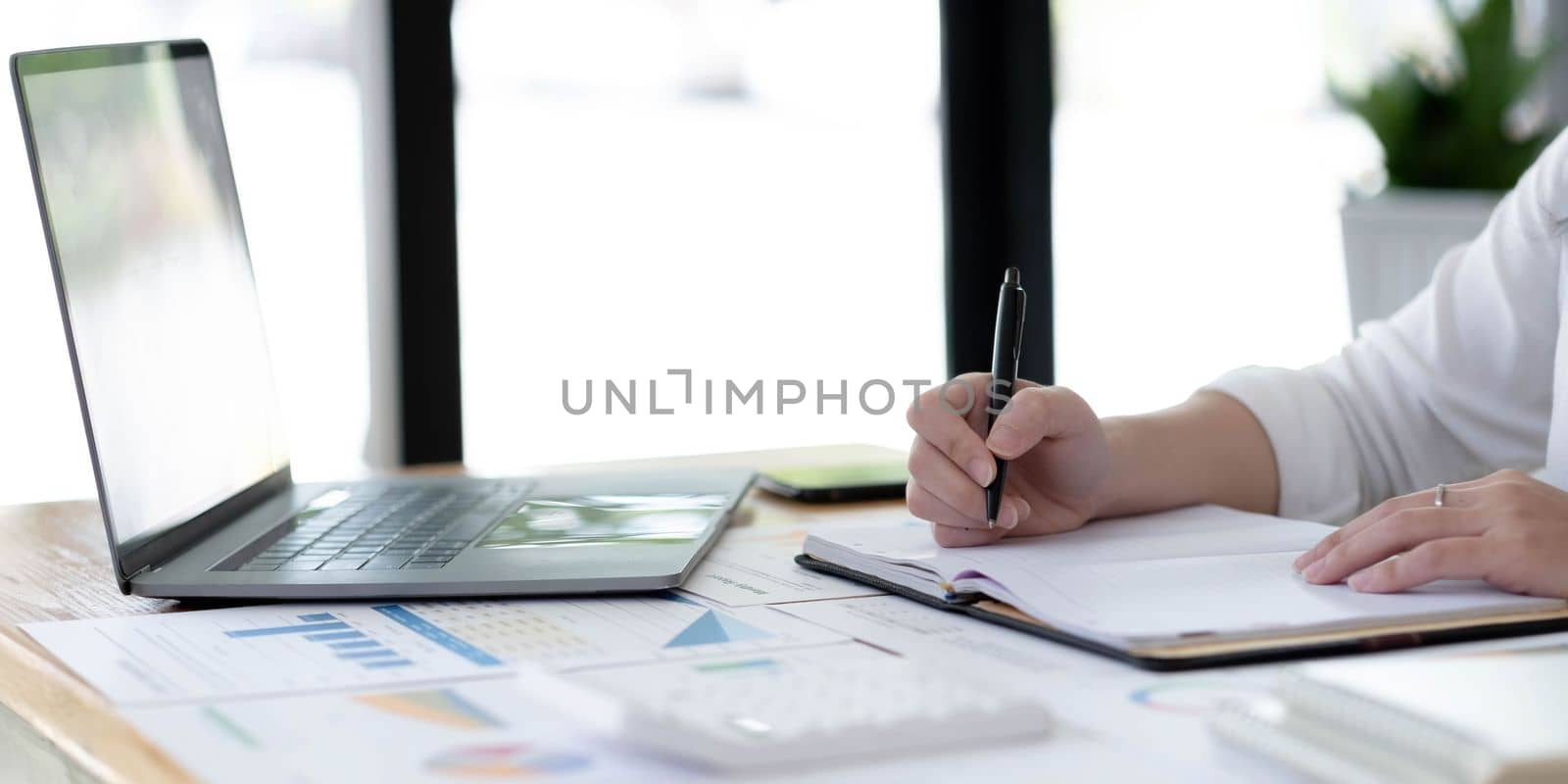 Young business woman sitting at table and taking notes in notebook.On table is laptop, smartphone by wichayada