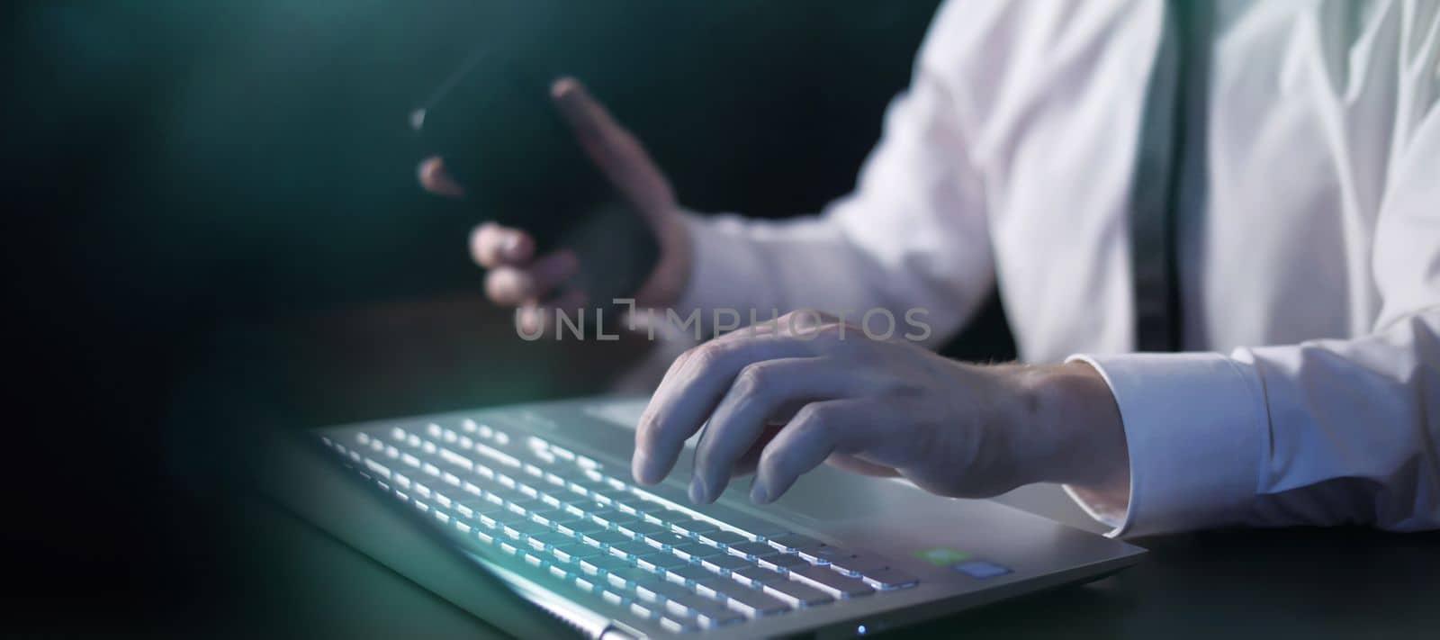 Businessman in white shirt working with a laptop while holding a mobile phone in his hand.