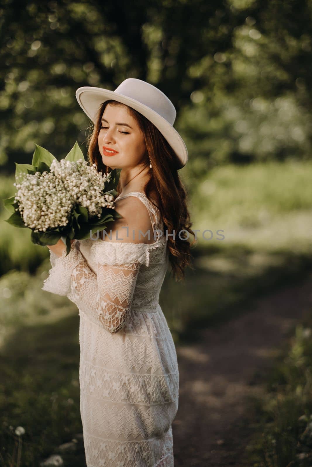 Portrait of a beautiful woman in a white dress and a hat with lilies of the valley. A girl in nature. Spring flowers by Lobachad