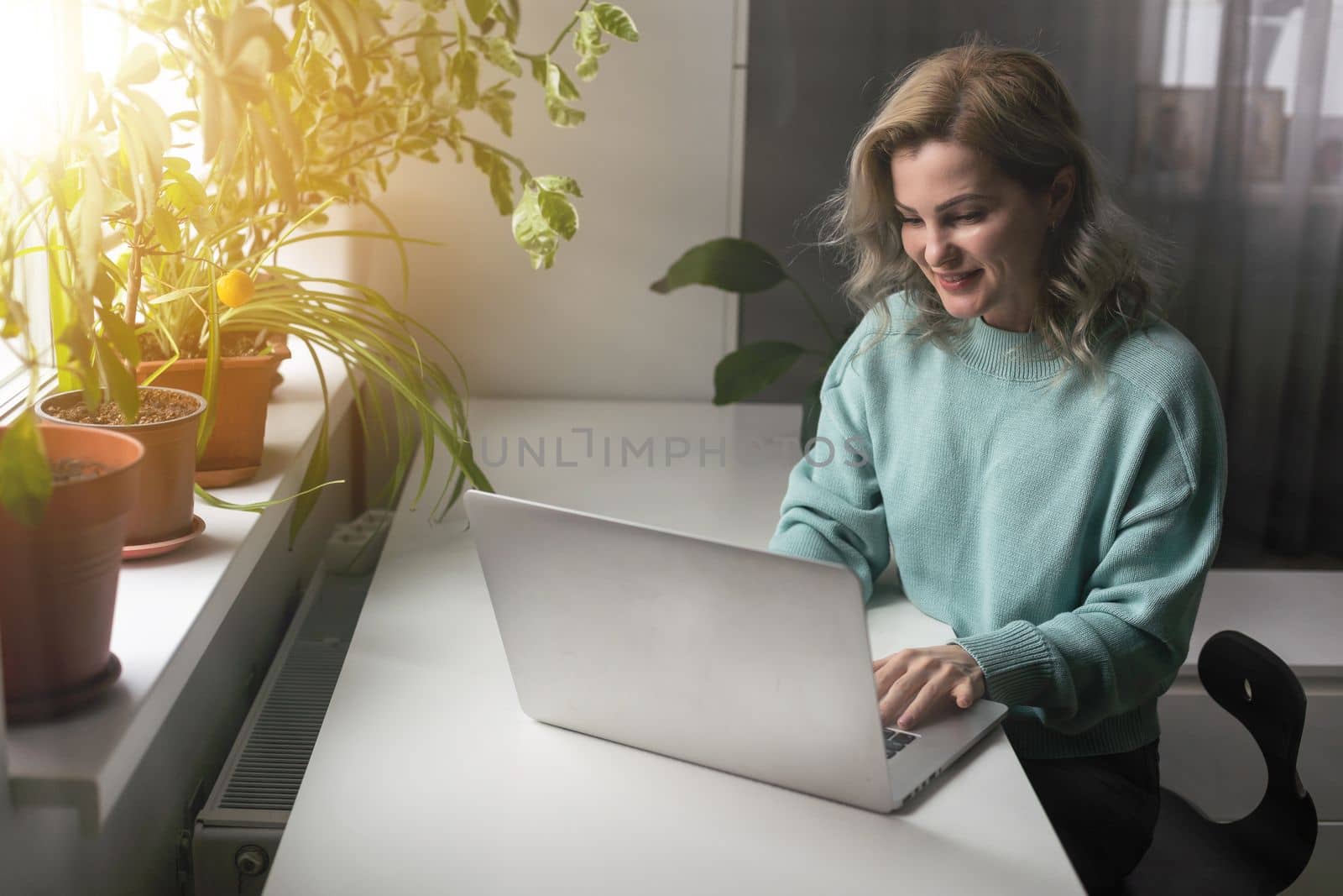 Positive young woman using a laptop at home, video conference. Cozy home interior with indoor plants. Remote work, business, freelance, online shopping, e-learning concept by Andelov13