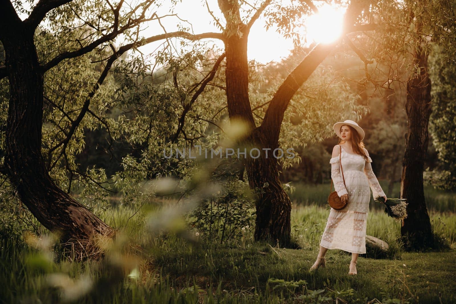 Portrait of a beautiful woman in a white dress and a hat with lilies of the valley at sunset. A girl in nature. Spring flowers by Lobachad