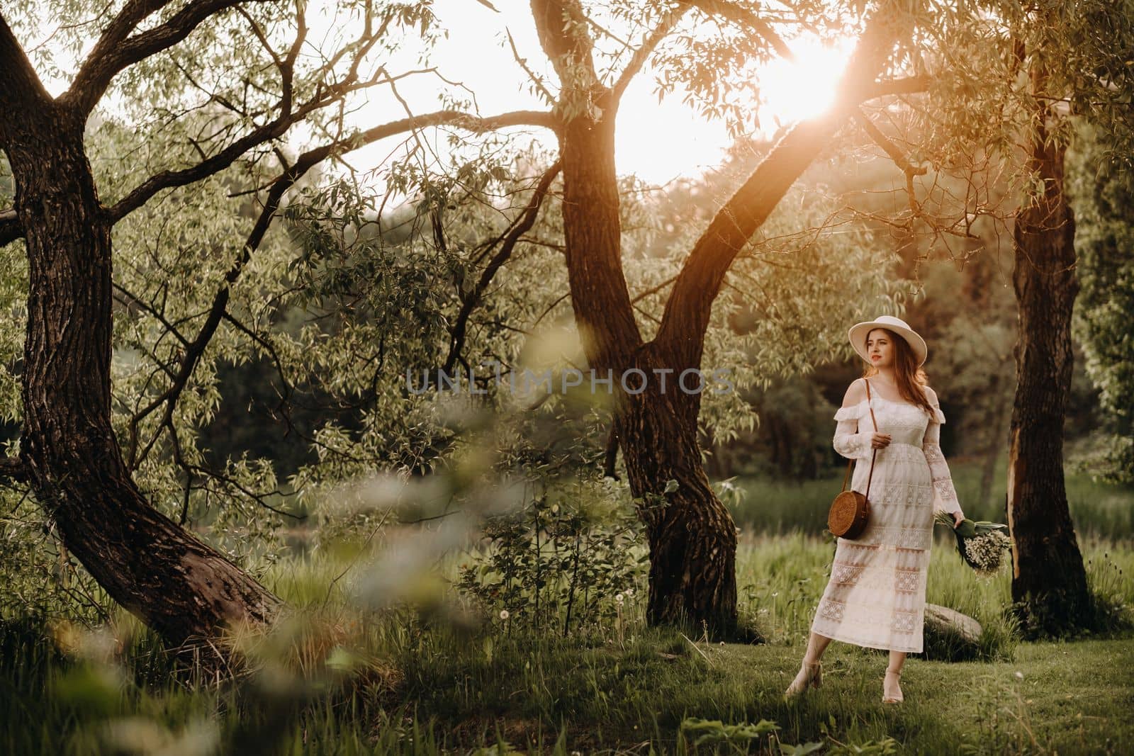Portrait of a beautiful woman in a white dress and a hat with lilies of the valley at sunset. A girl in nature. Spring flowers by Lobachad