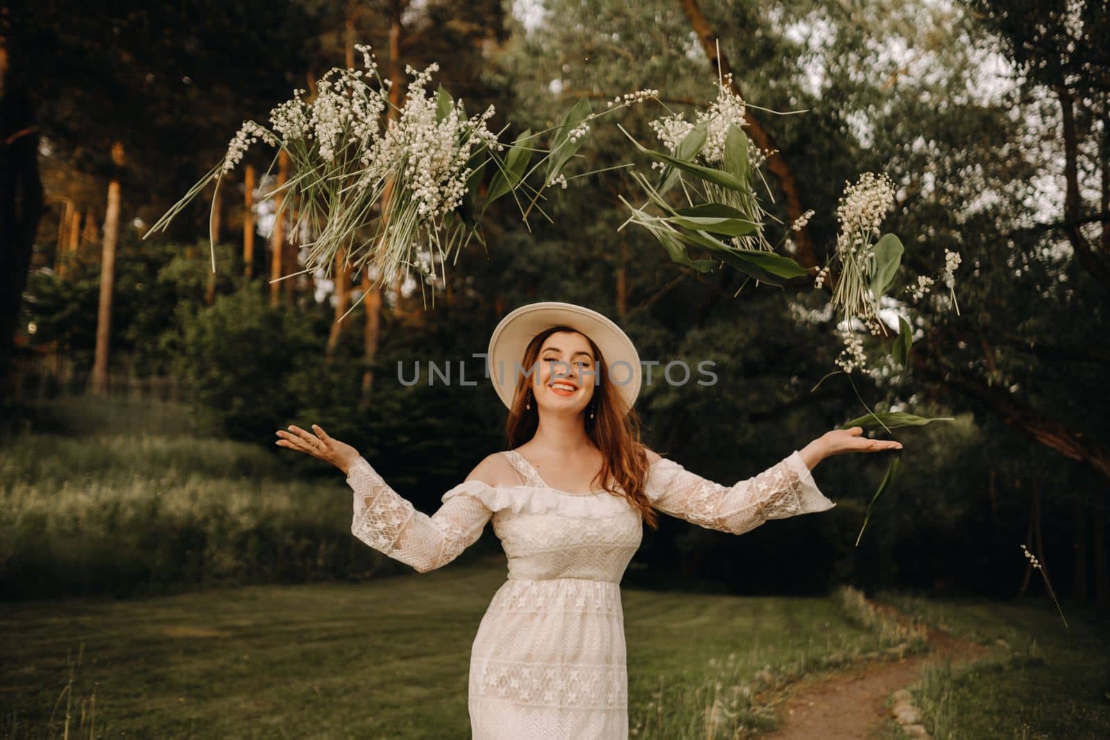 Portrait of a beautiful woman in a white dress and hat tossing lilies of the valley. A girl in nature. Spring flowers.