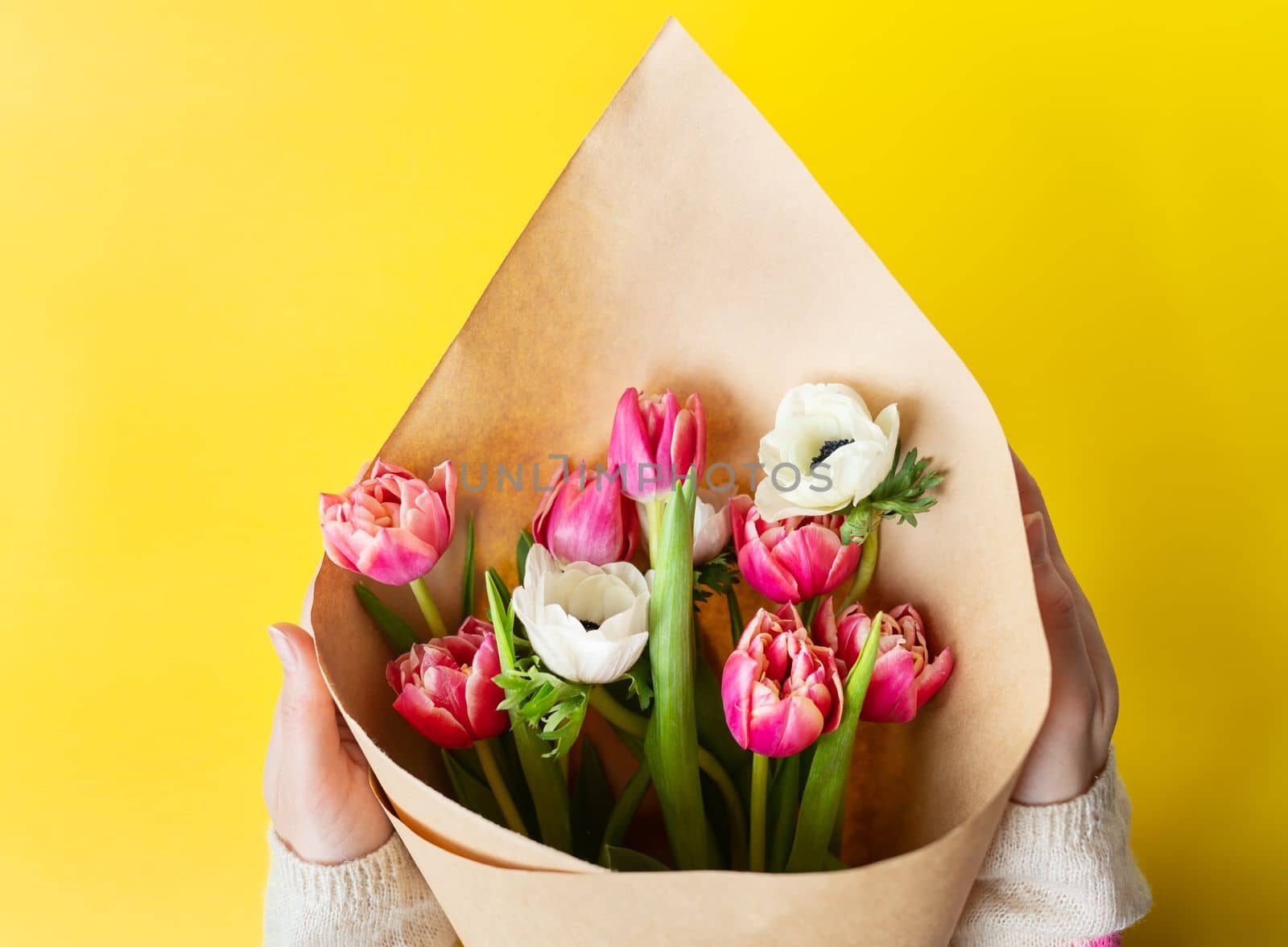 A girl holds a red-white bouquet of tulips and anemones in craft paper on a yellow background. With copy space