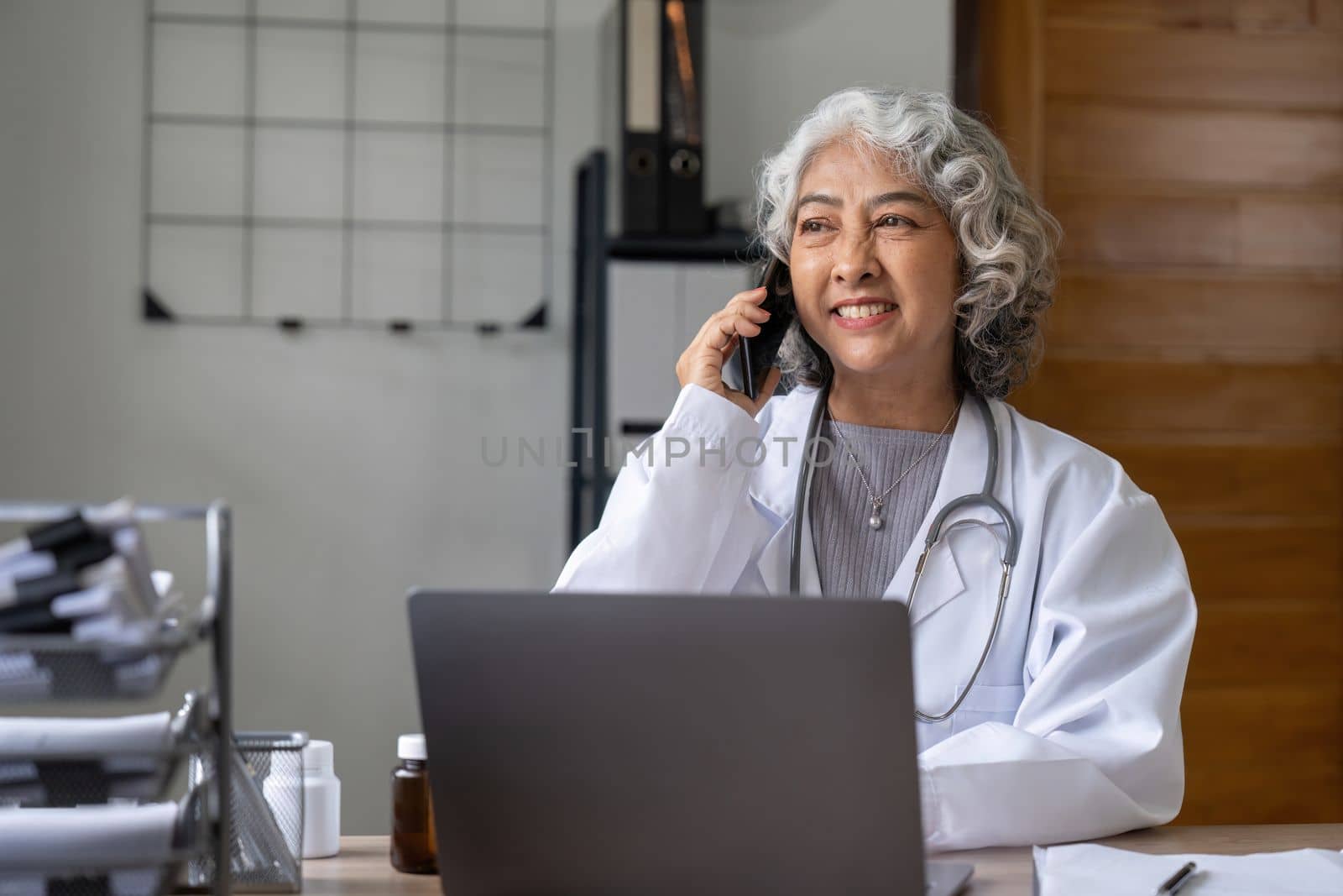 Senior asian female doctor using mobile phone talking while sitting in hospital office. happy woman medical worker having joyful conversation with husband in break time. nurse resting in clinic by nateemee