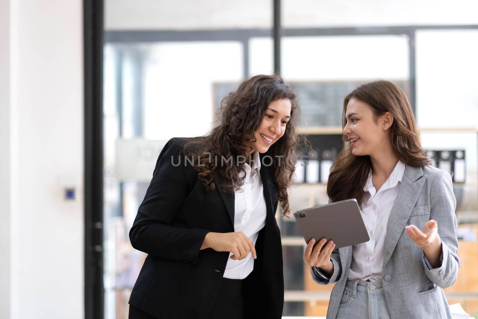 Two beautiful Asian businesswomen standing using digital tablet consulting and analyzing information in office work. by wichayada