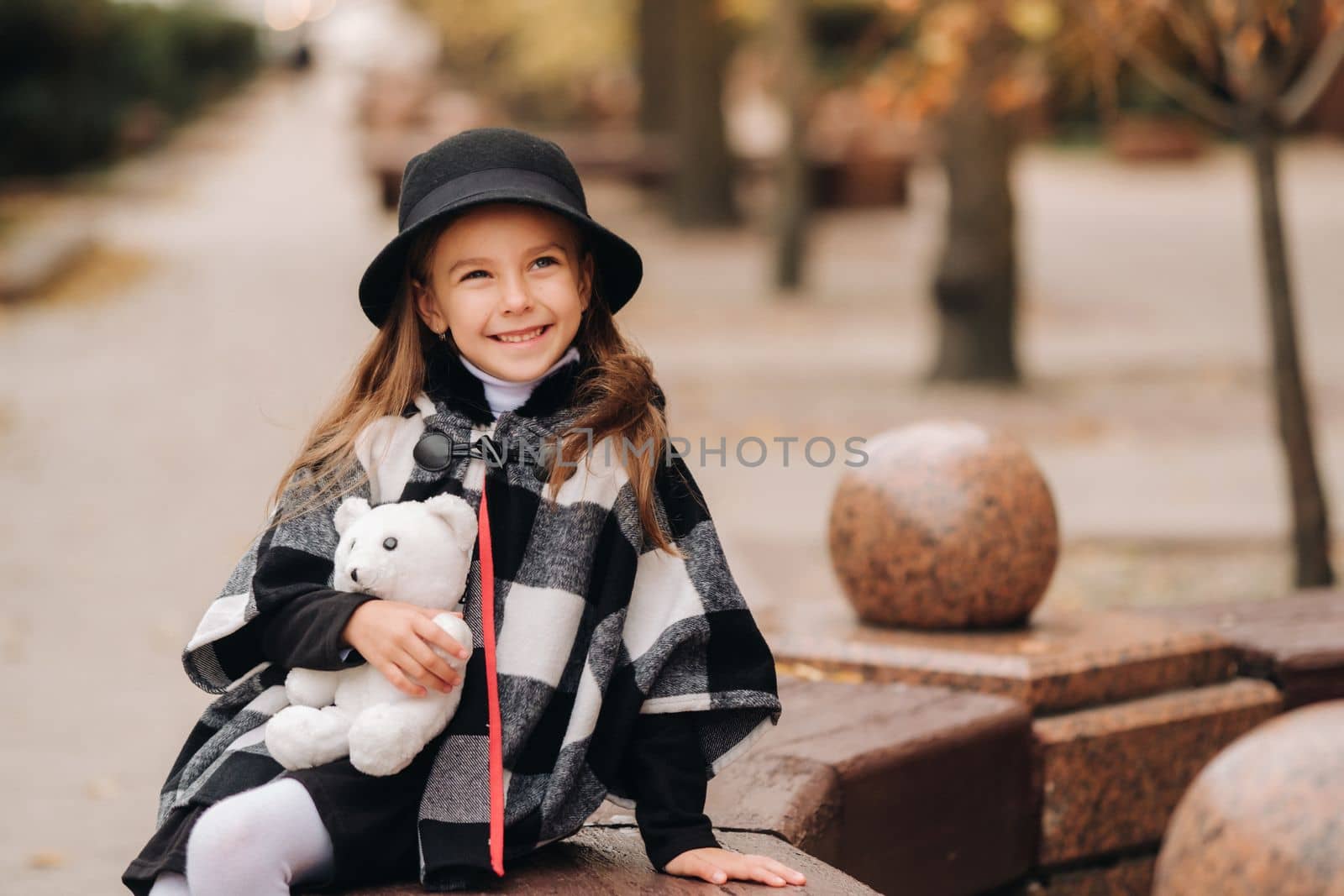 A little stylish girl in autumn clothes is sitting on a bench in the autumn city.