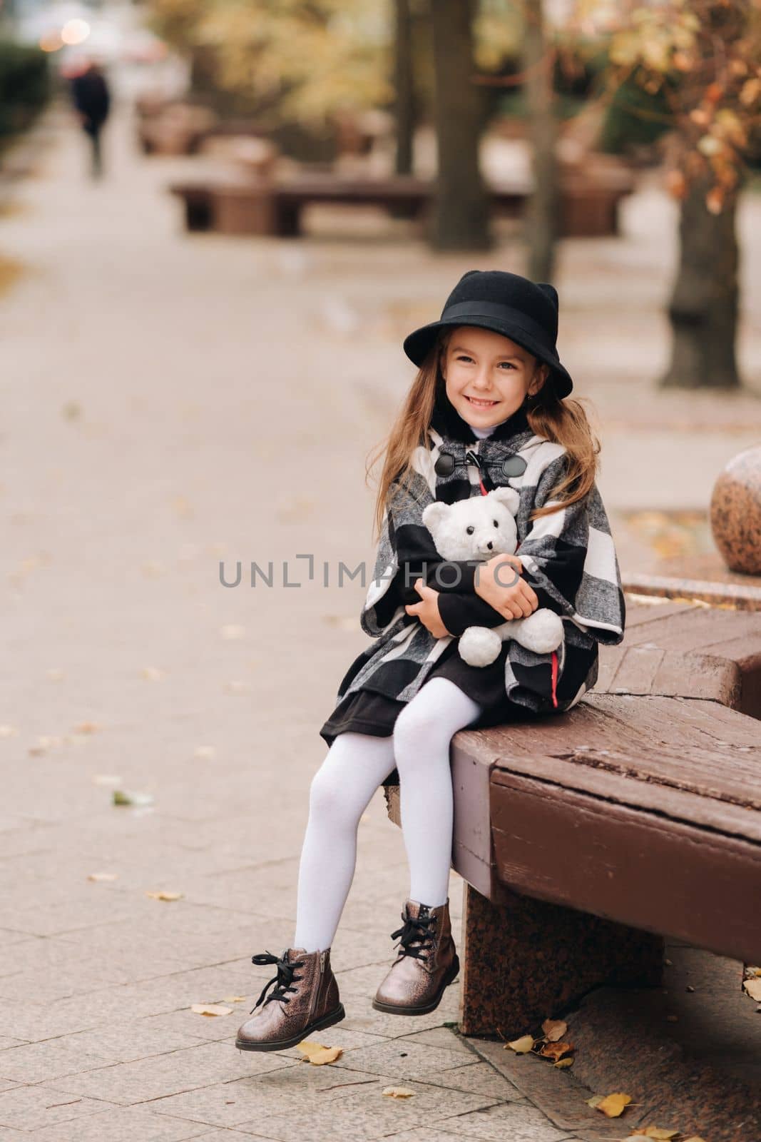 A little stylish girl in autumn clothes is sitting on a bench in the autumn city by Lobachad