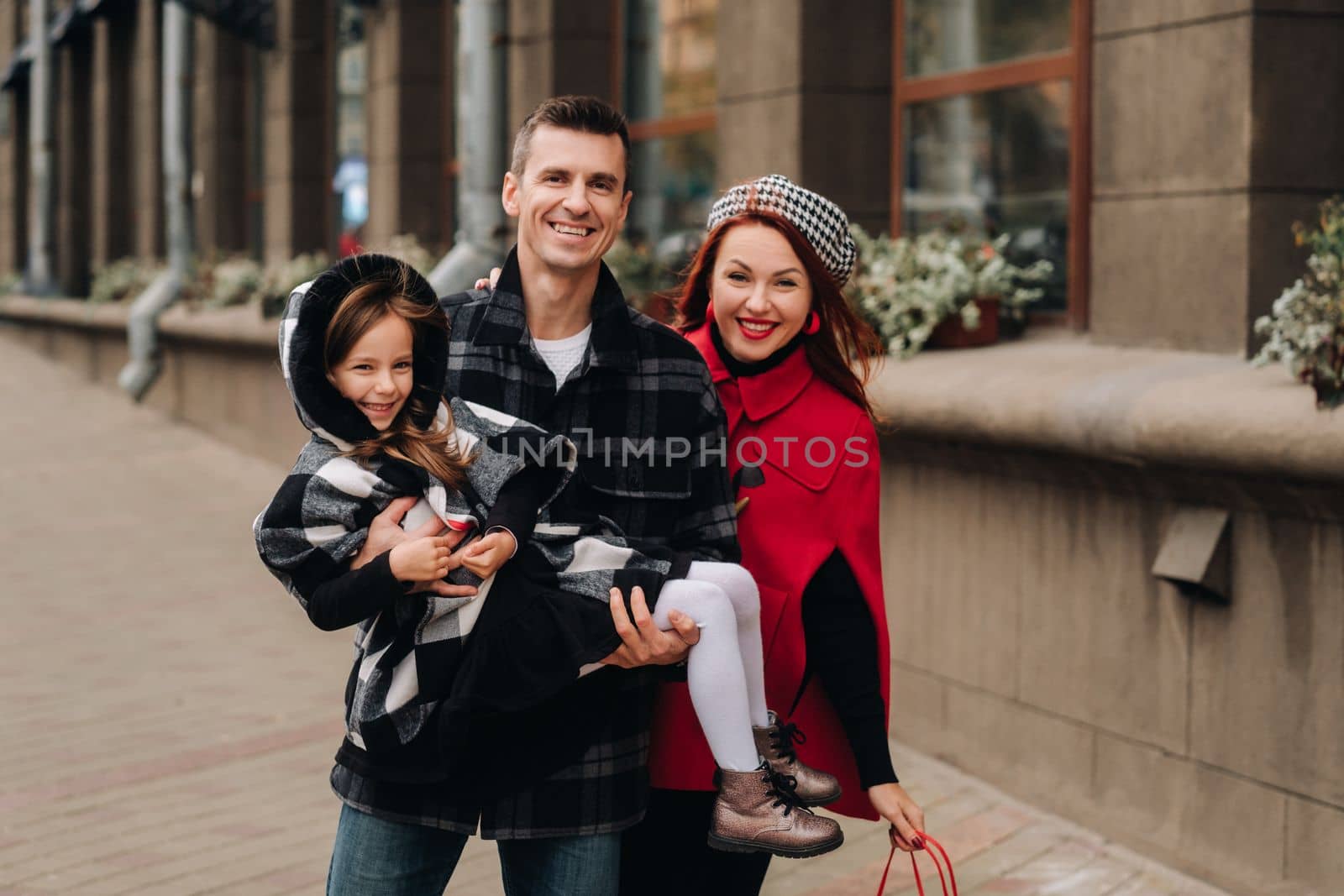 A stylish family of three people are walking in the city in the fall and dad is holding his daughter in his arms by Lobachad