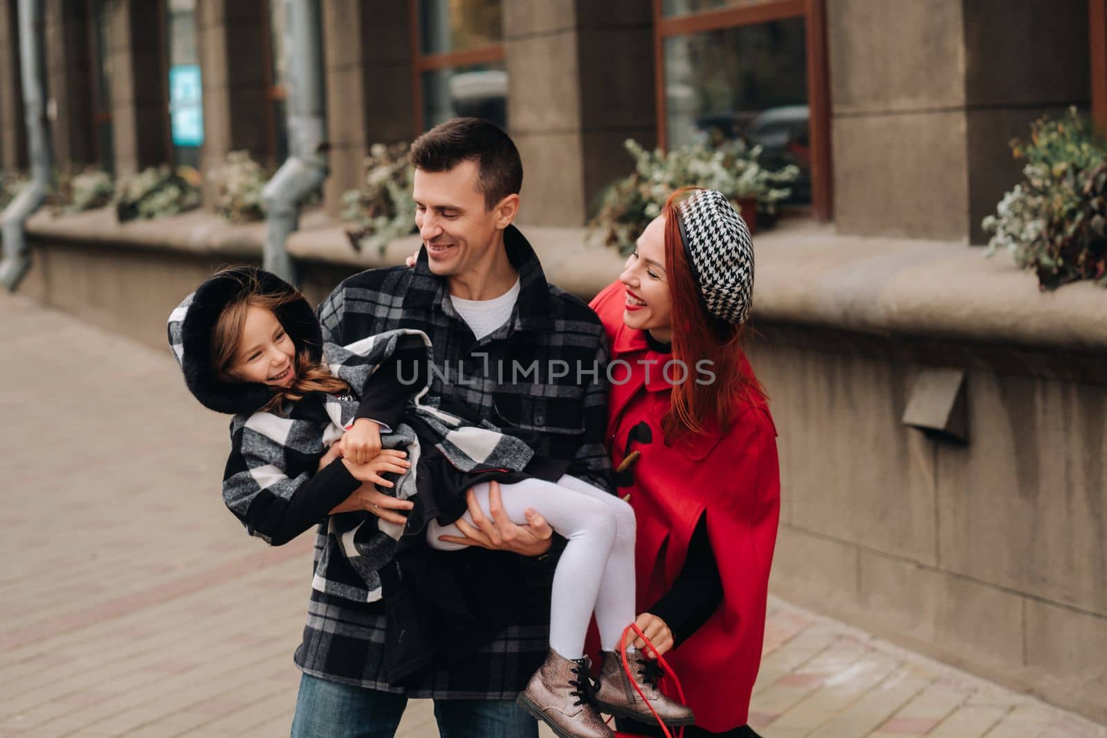 A stylish family of three people are walking in the city in the fall and dad is holding his daughter in his arms.