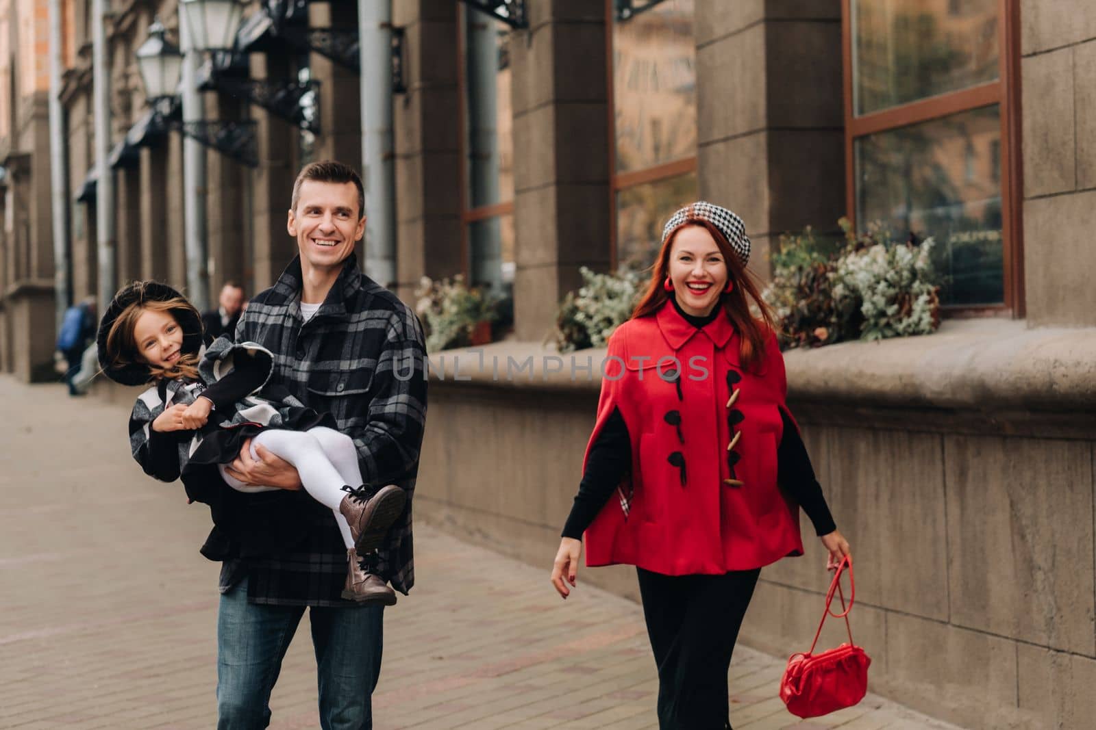 A stylish family of three people are walking in the city in the fall and dad is holding his daughter in his arms.