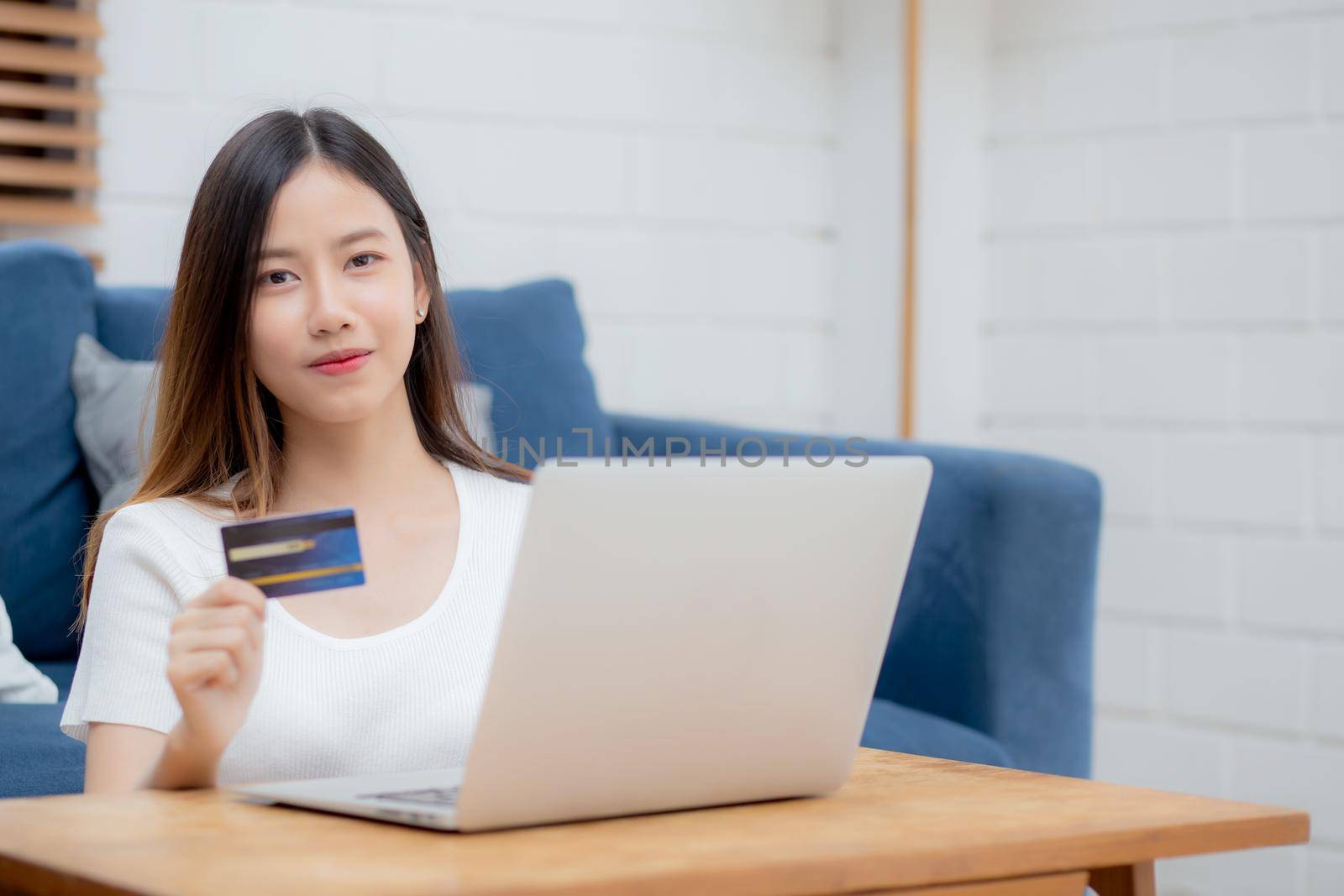 Young asian woman sit on sofa using laptop computer shopping online with credit card buying to internet, happy girl payment with e-business on couch, purchase and payment, business concept. by nnudoo