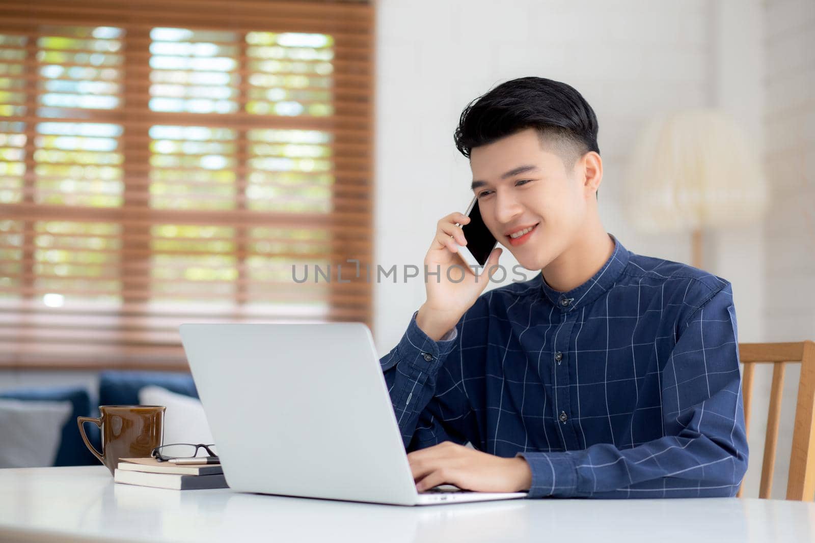 Young asian man talking phone and work from home with laptop computer, freelance using notebook to internet online and speak on smartphone, domestic life, business and communication concept. by nnudoo