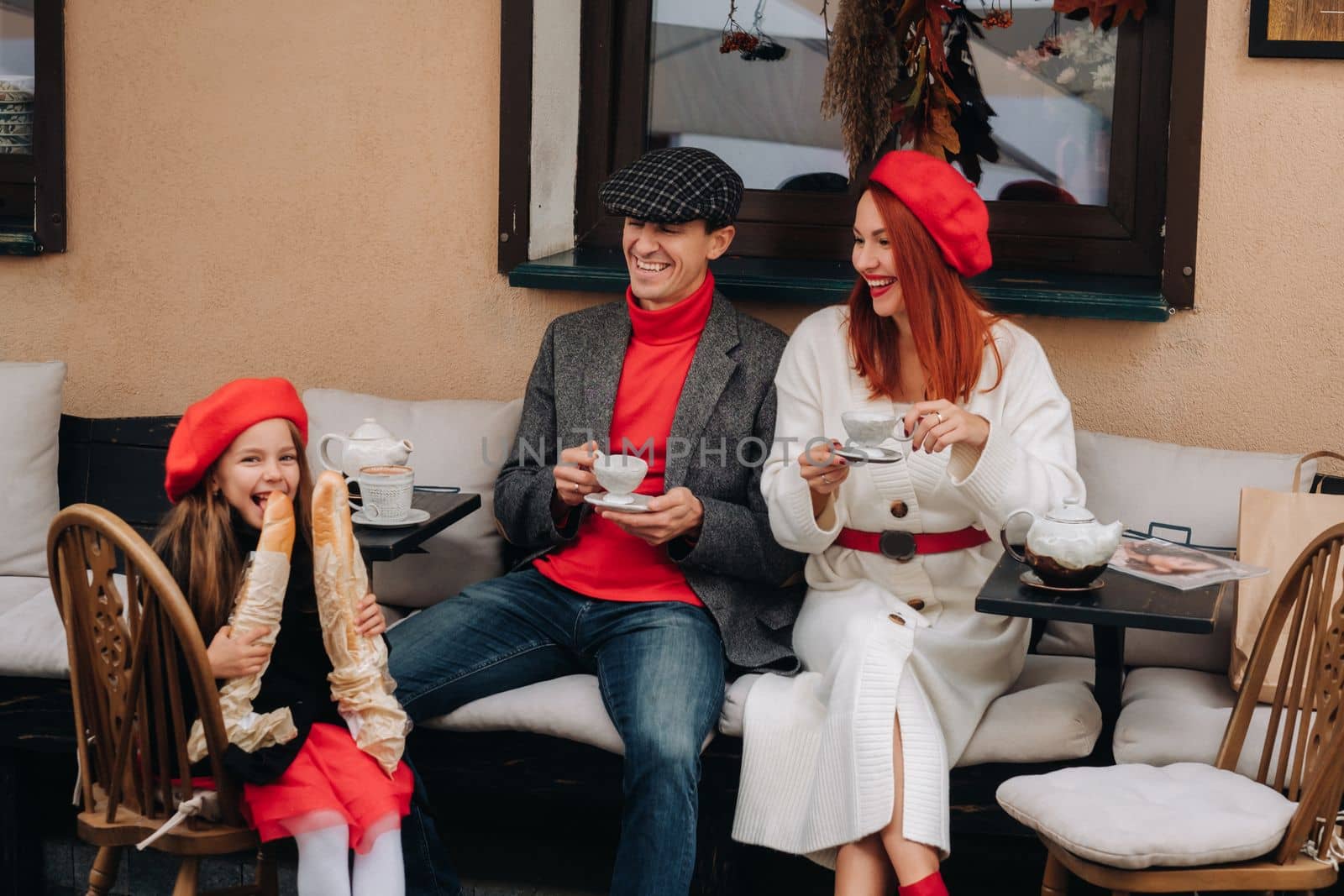 A stylish family of three is sitting at a table outside in a cafe and drinking coffee. Dad, mom and daughter in the autumn city.