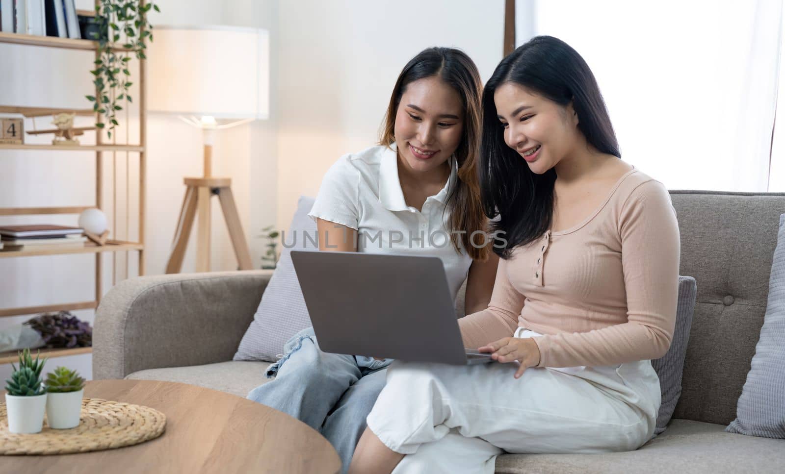 young asian women LGBT lesbian happy couple sitting on sofa using laptop a computer and phone in living room at home. LGBT lesbian couple together