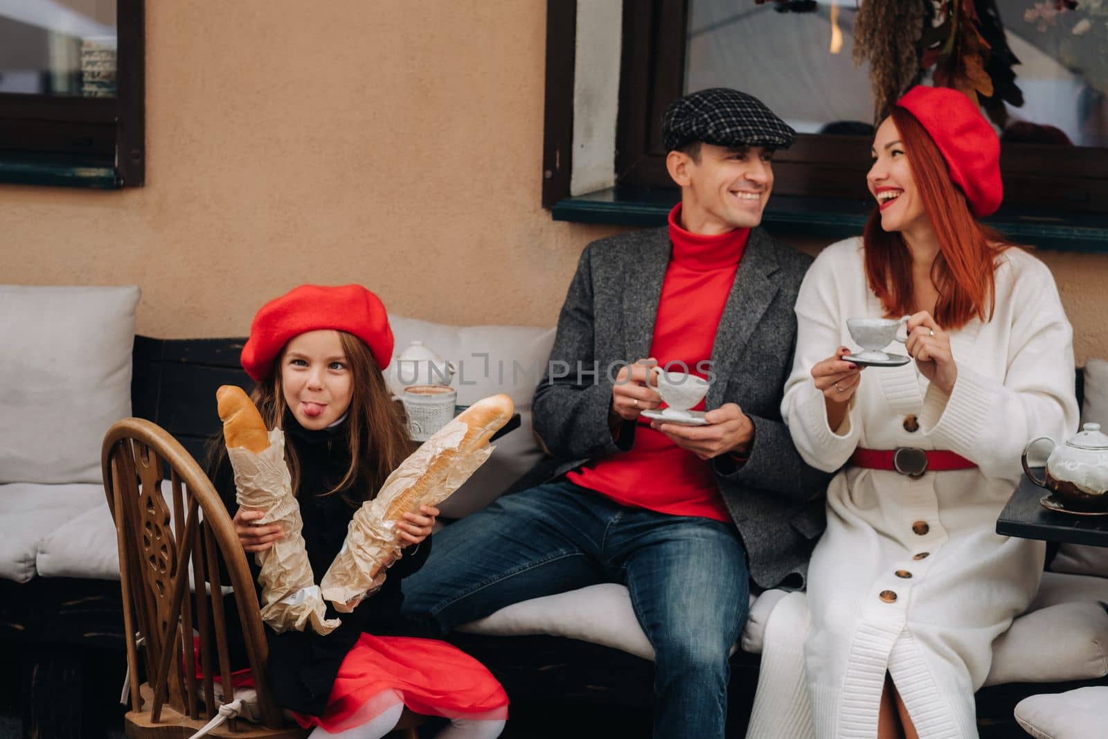 A stylish family of three is sitting at a table outside in a cafe and drinking coffee. Dad, mom and daughter in the autumn city.