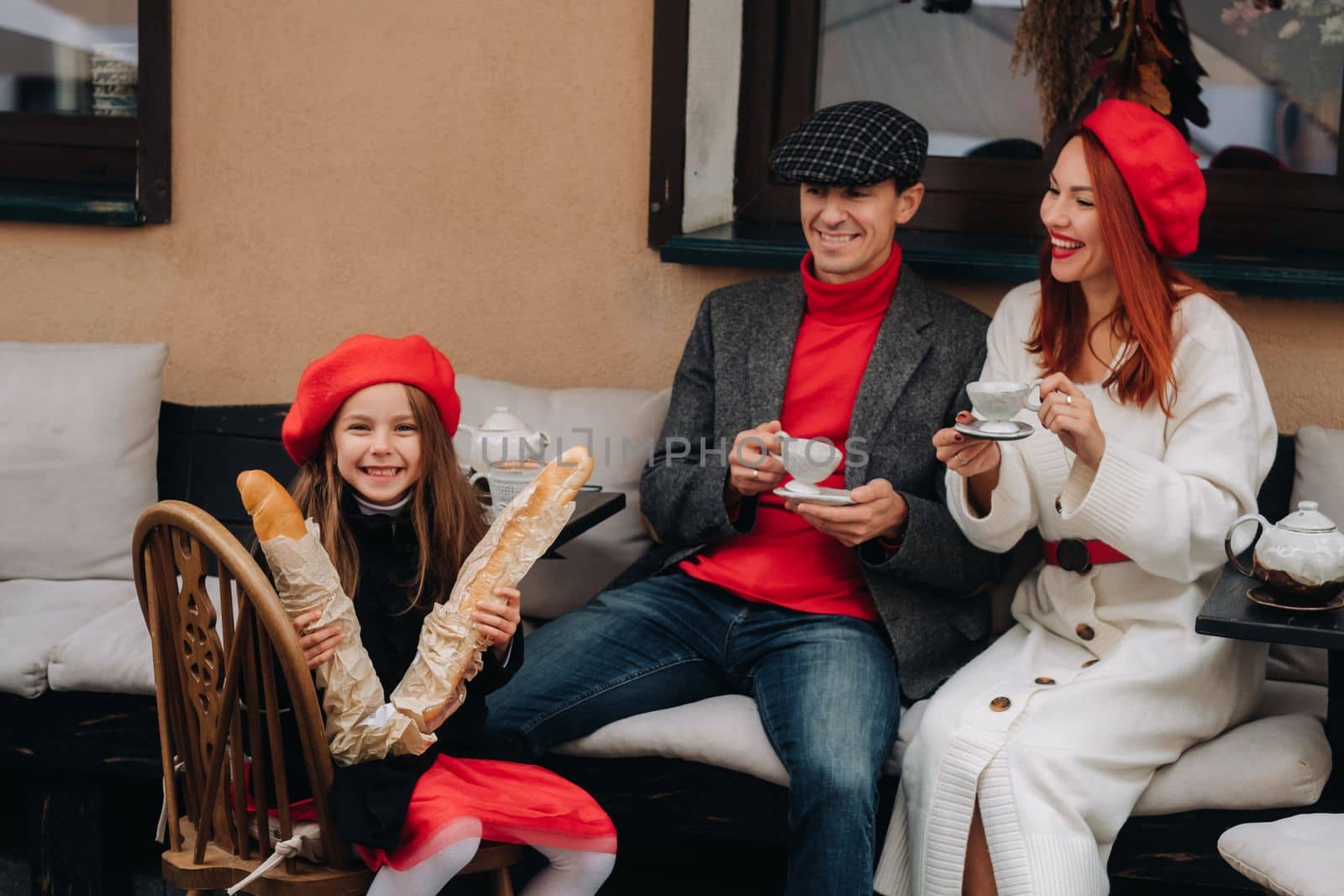 A stylish family of three is sitting at a table outside in a cafe and drinking coffee. Dad, mom and daughter in the autumn city by Lobachad