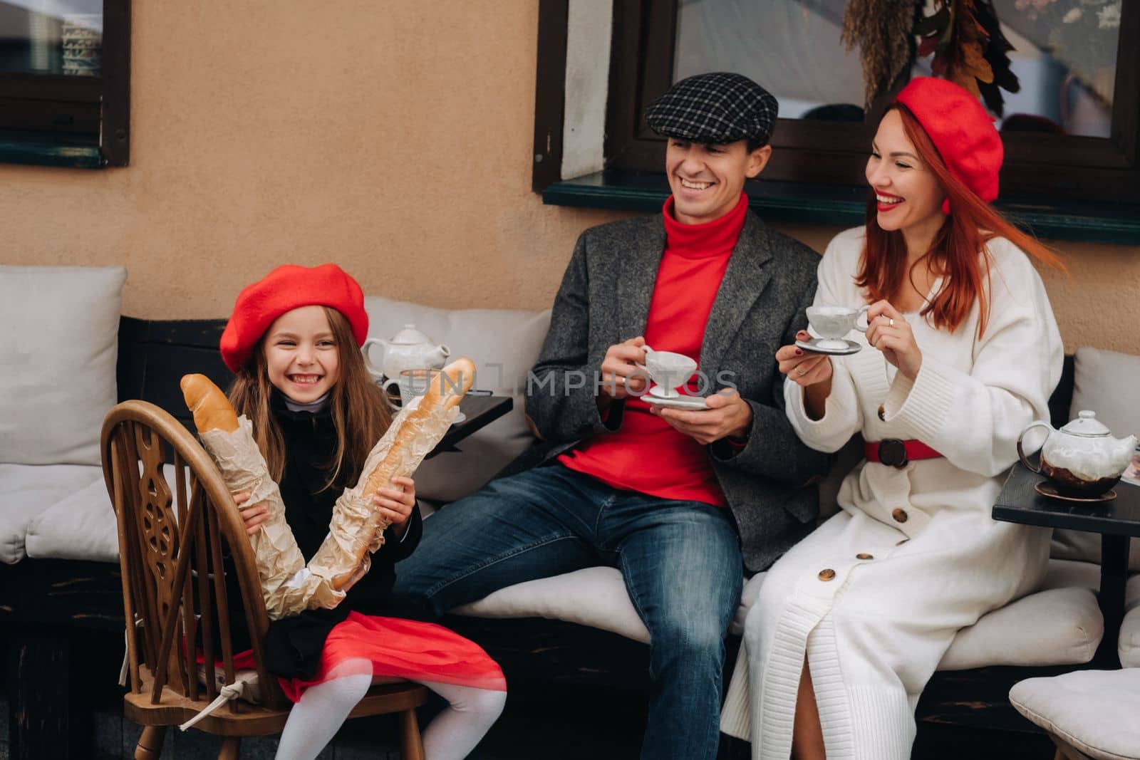 A stylish family of three is sitting at a table outside in a cafe and drinking coffee. Dad, mom and daughter in the autumn city.