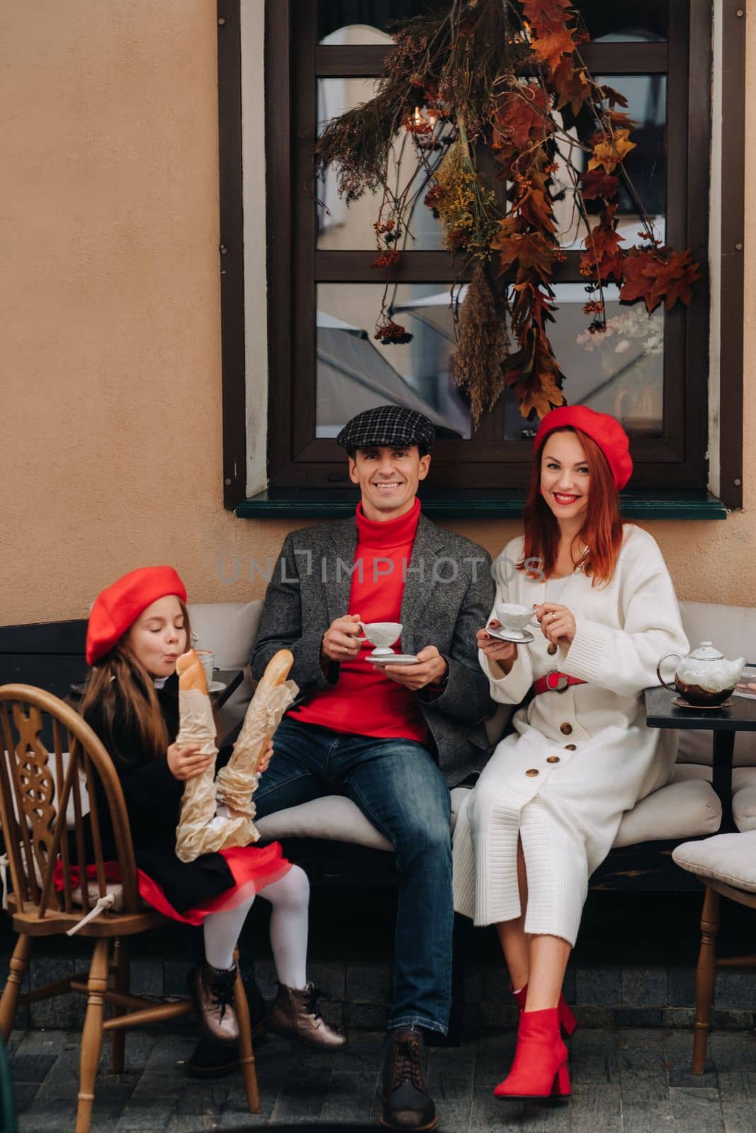 A stylish family of three is sitting at a table outside in a cafe and drinking coffee. Dad, mom and daughter in the autumn city.