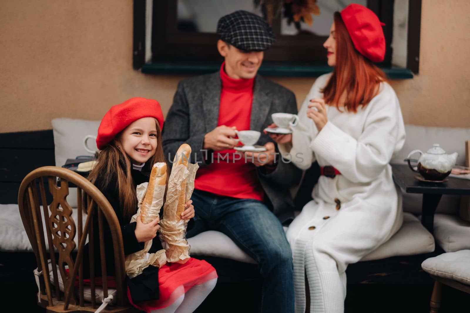 A stylish family of three is sitting at a table outside in a cafe and drinking coffee. Dad, mom and daughter in the autumn city by Lobachad