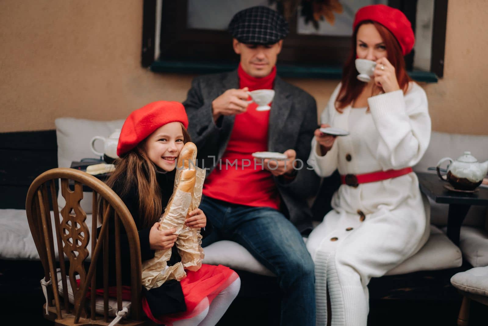 A stylish family of three is sitting at a table outside in a cafe and drinking coffee. Dad, mom and daughter in the autumn city.