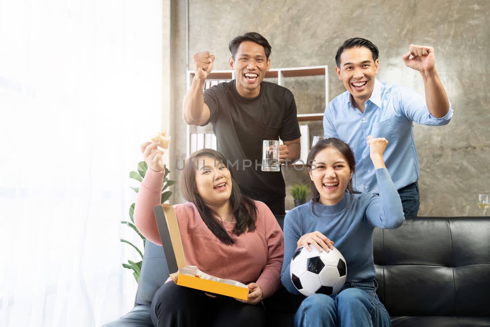 Friends watch sports on TV, cheer and celebrate. Happy diverse asian friend supporters fans sit on couch with popcorn and drinks.